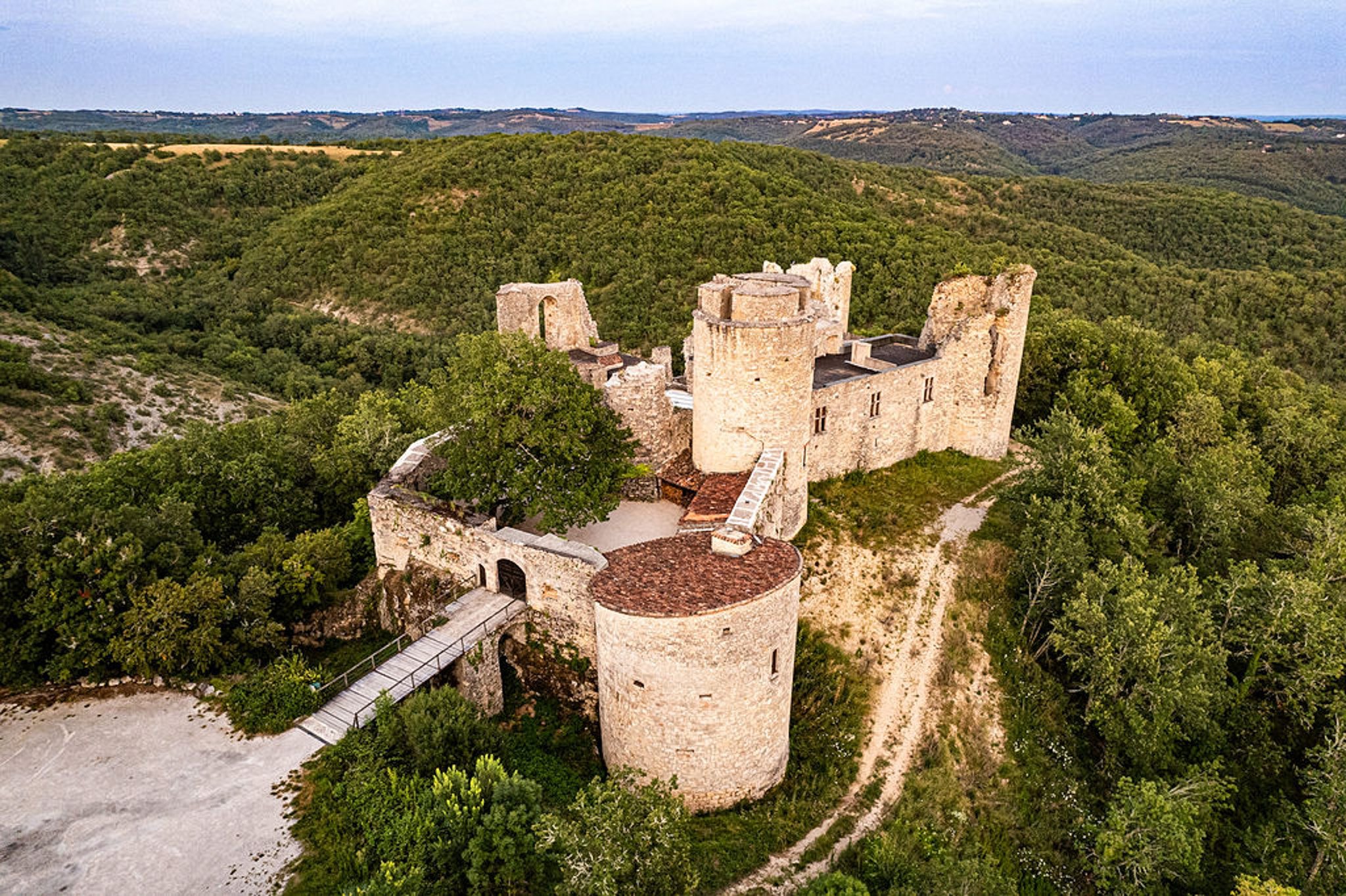 Aerial drone shot of a medieval French castle ruin set atop a hillside, surrounded by dense woodland stretching across rolling hills to the horizon. The structure features multiple round towers in pale limestone, partially intact battlements, and a large cylindrical keep with a terracotta-tiled roof in the foreground. A wooden footbridge with metal railings connects the entrance gateway to a gravel parking area at lower left. No people, wedding party, or event decor are visible in this image. Potential venue feature image.