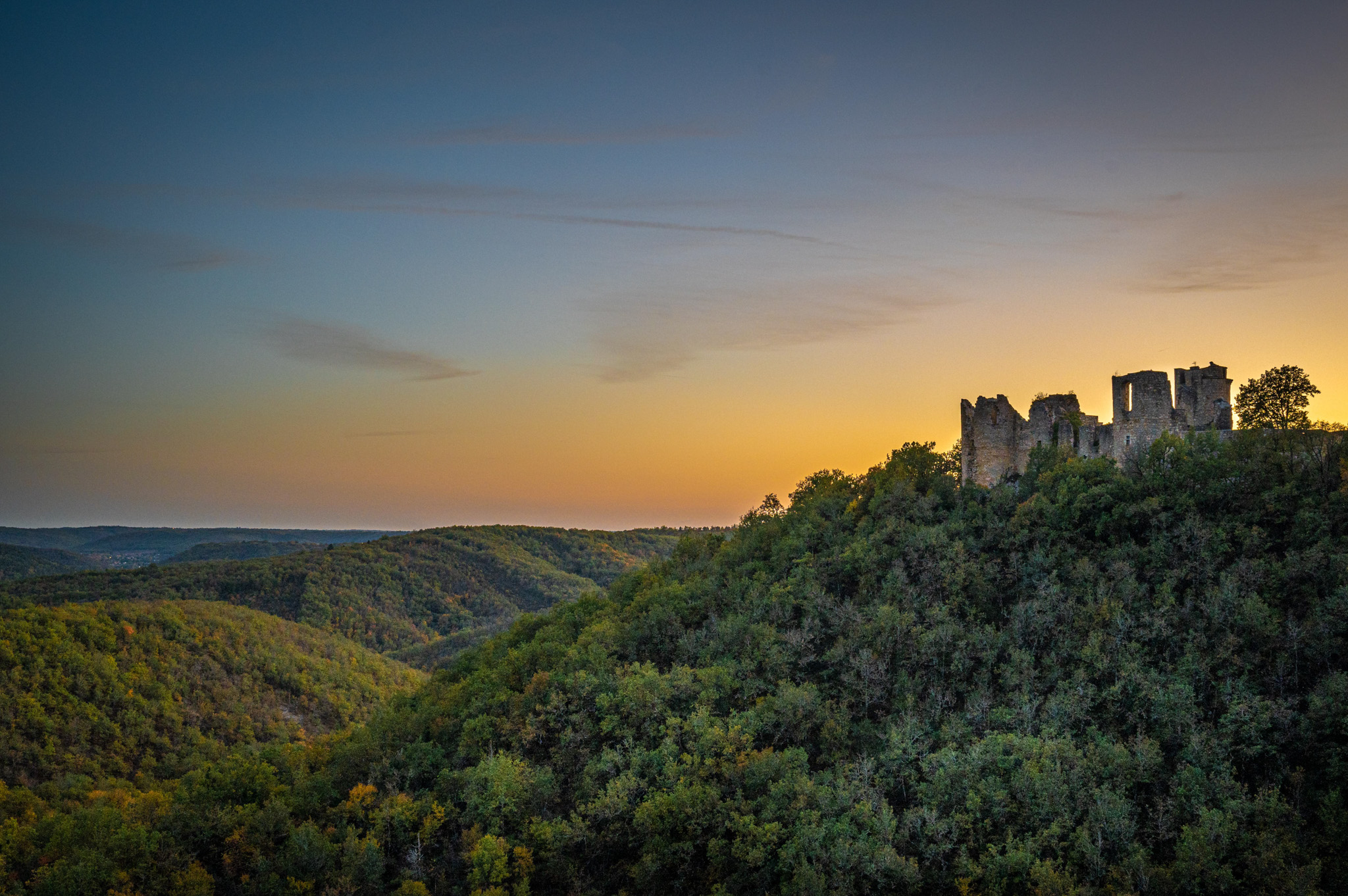 Wide-angle aerial or elevated landscape shot showing the ruins of a medieval stone castle or fortification perched on a densely forested hilltop, with rolling wooded valleys extending into the distance. No people, wedding party, or wedding-related decor are visible in this image — it appears to be a standalone venue or location establishing shot taken at dusk, with a warm amber and golden horizon transitioning to a blue-grey upper sky. The castle ruins feature multiple remaining stone towers and walls in partial collapse. Potential venue feature image.