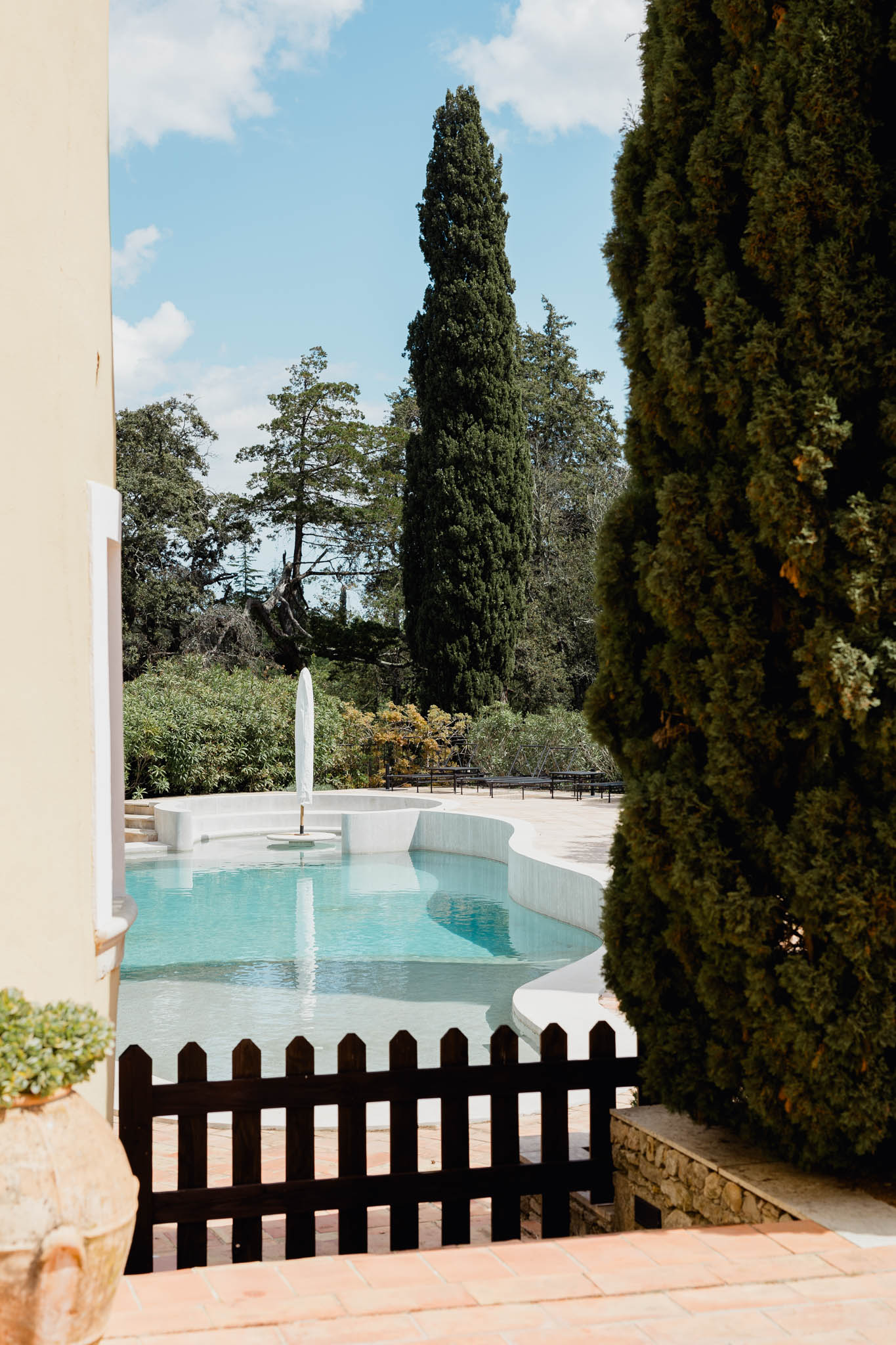 Freeform pool with fountain feature viewed through gap between cream building and cypress tree at French venue