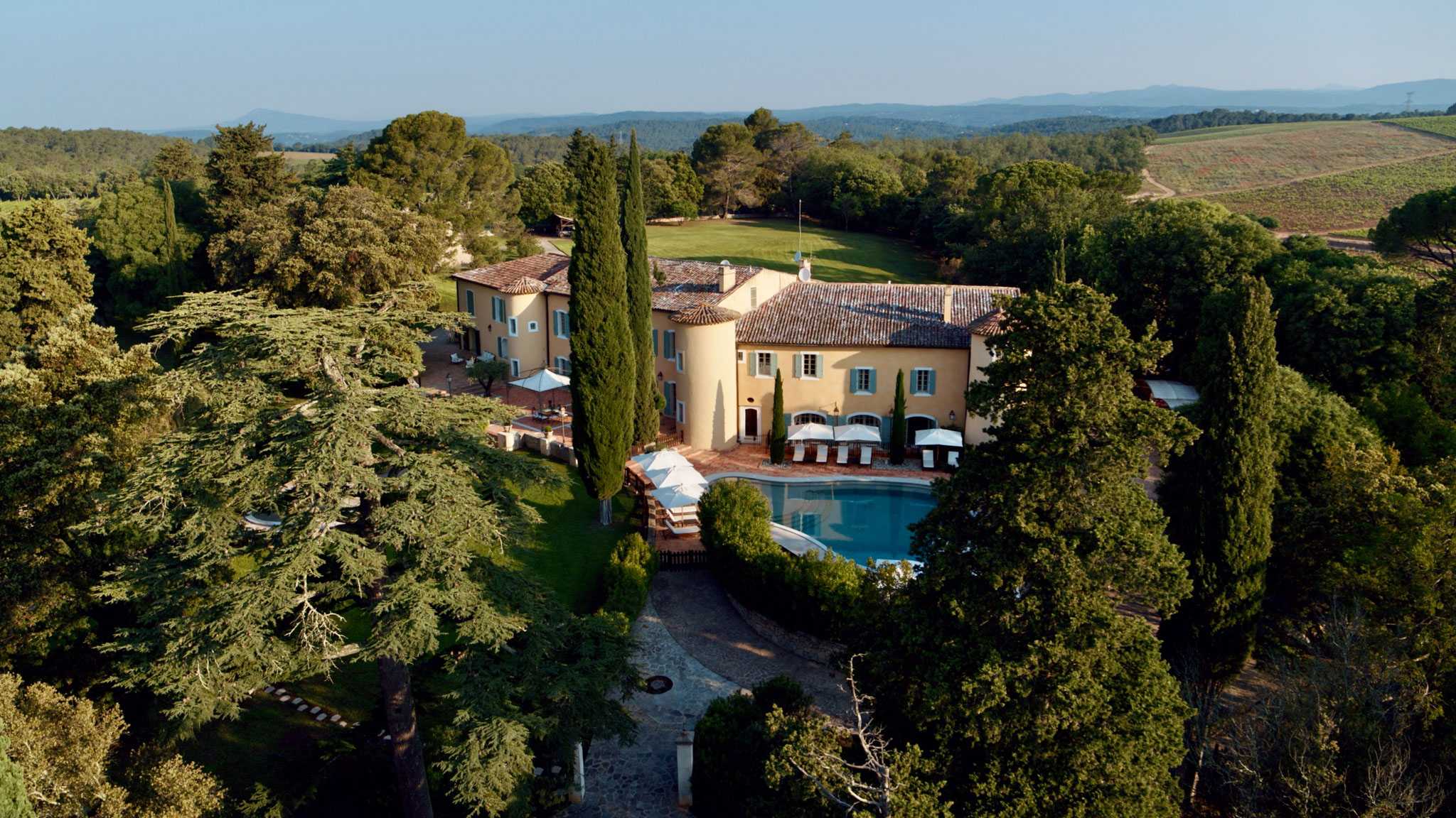 Aerial view of Provencal chateau with yellow facades, swimming pool, cypress trees, and vineyard backdrop