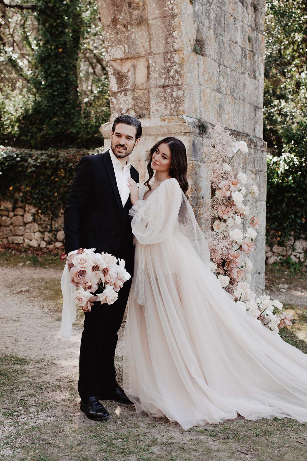 A couple portrait taken outdoors against an ancient stone column or ruin, with ivy-covered walls visible in the background. The bride wears a blush-toned tulle gown with billowing long sleeves and a flowing skirt, and holds a rounded bouquet of blush garden roses, deep mauve roses, and white blooms with trailing white ribbon. The groom wears a black suit with a white dress shirt and no tie. Behind them, a floral installation cascades along the stone column featuring blush and white roses, orchids, and baby's breath. The styling leans toward romantic and organic, with a soft blush and white color palette. This is a mid-length couple portrait shot.