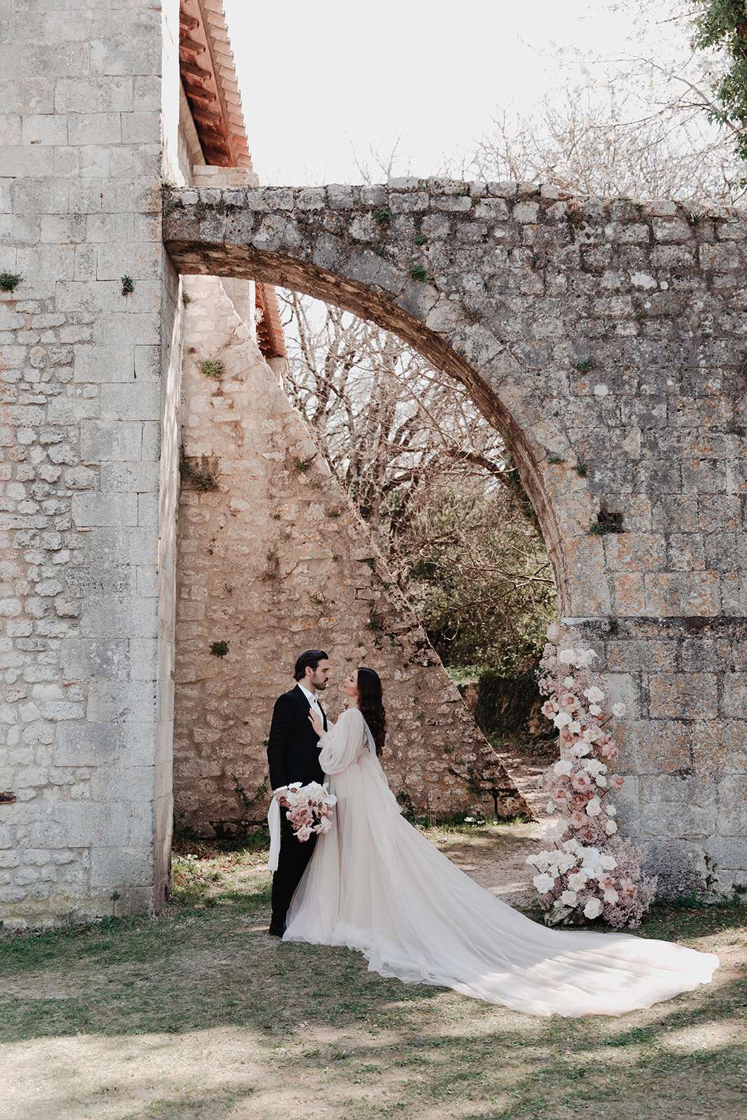 A couple portrait taken outdoors at the base of a large stone arch, part of what appears to be a historic ruined structure or chapel exterior. The bride wears a soft white tulle ballgown with long billowing sleeves and a very long cathedral-length train spread across the ground; she holds a bouquet of blush pink and white blooms including roses and orchids with trailing ribbons. The groom wears a black tuxedo with a white dress shirt and bow tie. To the right of the couple, a tall freestanding floral column is composed of white and blush pink roses, garden roses, and dried or tonal pink accent flowers. The couple faces each other in a close, intimate pose in this medium full-length portrait shot. The overall styling palette is blush, white, and black with a classic romantic aesthetic.