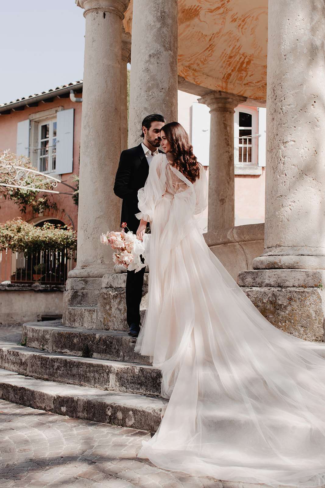 A couple portrait taken outdoors on the stone steps of a classical columned structure in what appears to be a Provençal village setting, with a terracotta-rendered building with pale blue shutters visible in the background. The bride wears a blush-to-ivory tulle gown with dramatic billowing long sleeves and an extended train that cascades down the steps, while the groom is dressed in a black tuxedo with a white shirt. The bride holds a loose, downward-facing bouquet of blush and pale pink peonies or garden roses. The two are posed close together, foreheads nearly touching, in a three-quarter full-length portrait shot that highlights the volume and length of the gown's train against the worn stone steps and cobblestone pavement.