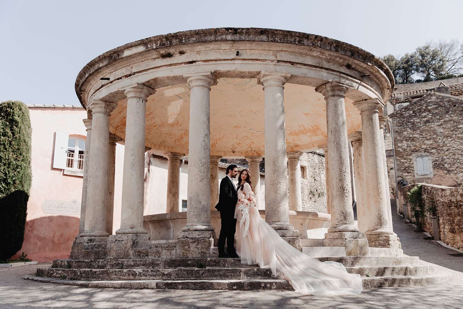 Bride in blush tulle gown with trailing train and groom at stone rotunda in Provencal village