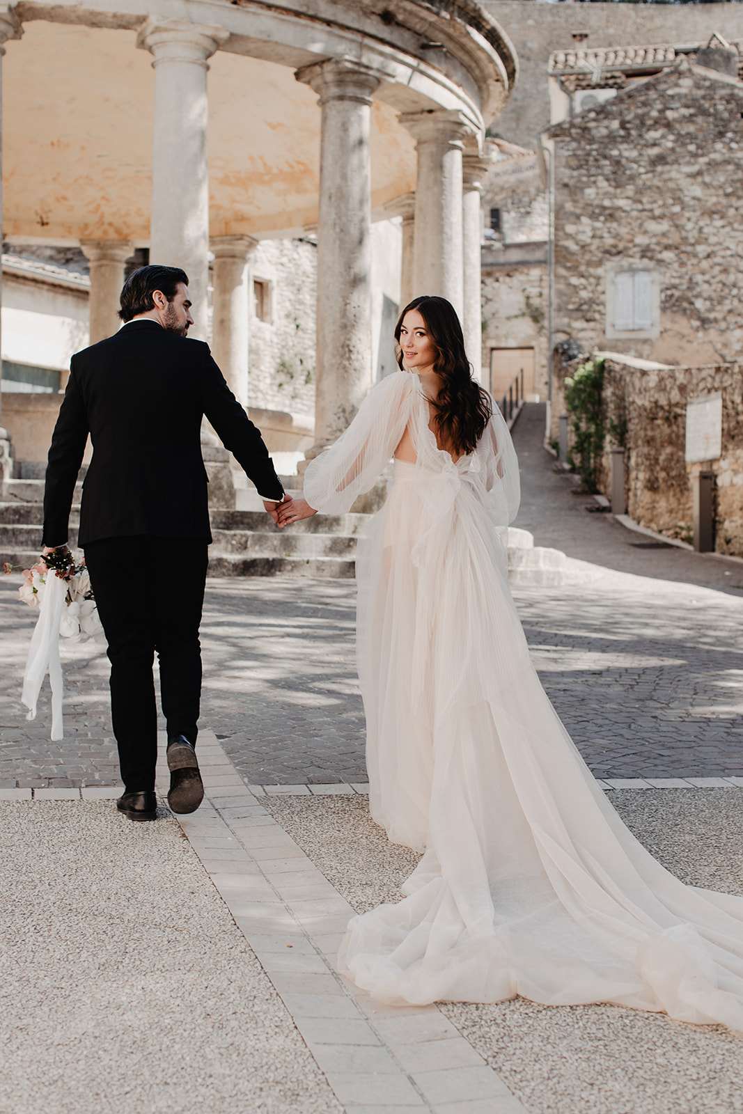 Bride and groom walking hand-in-hand across cobblestones in a southern French village square with stone colonnade