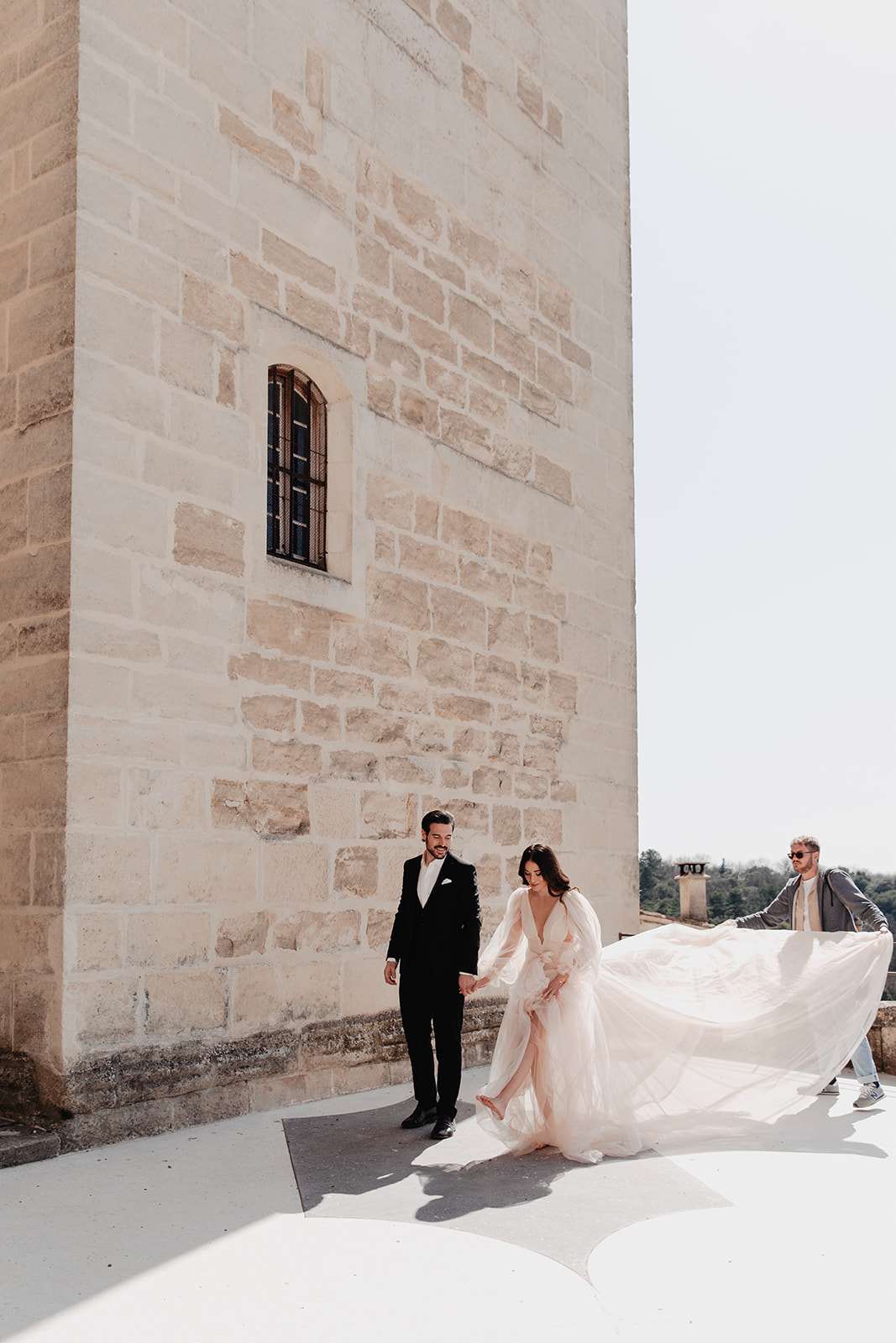 Bride in blush tulle gown with dramatic dress toss beside groom in black tuxedo on chateau rooftop
