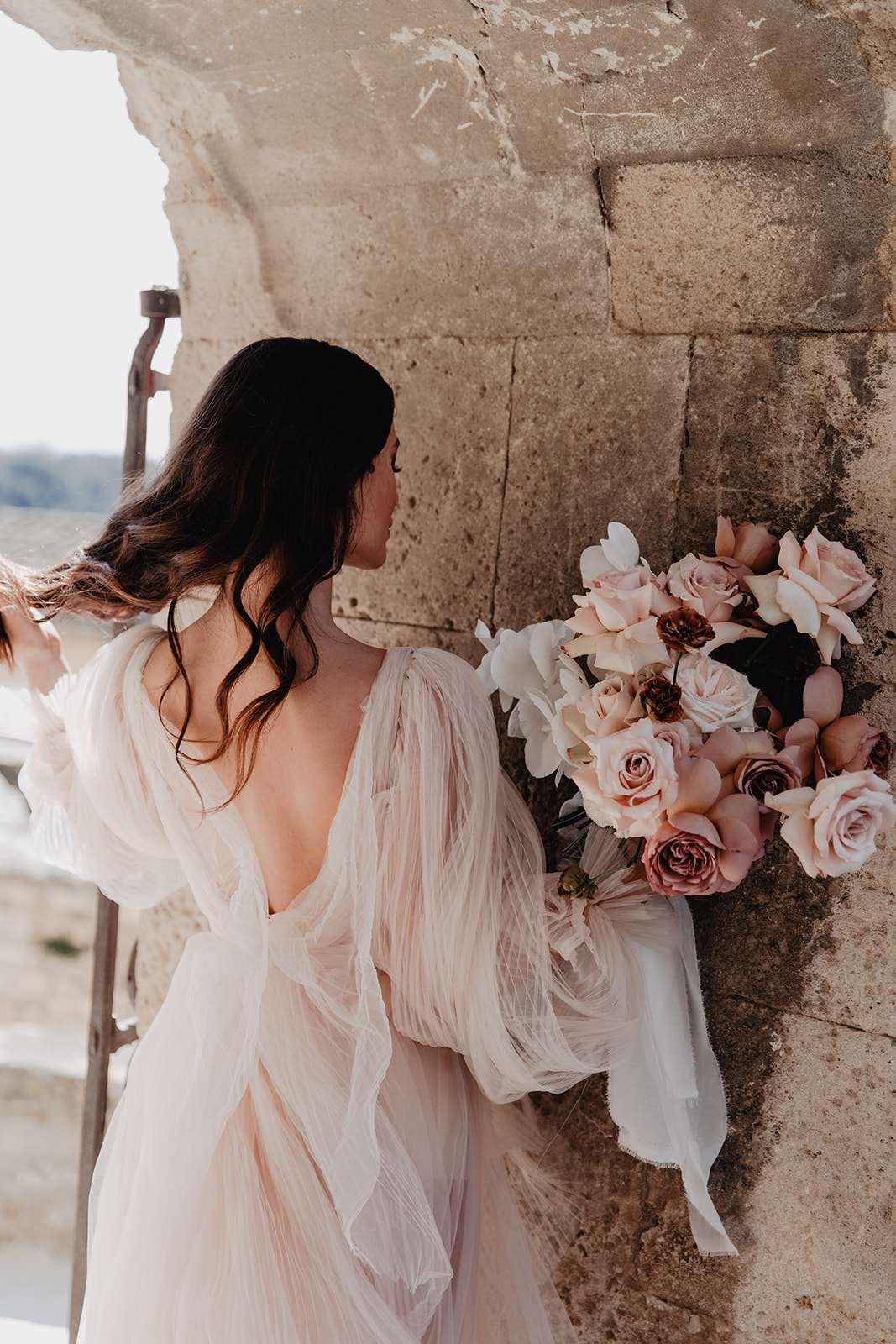 A bridal portrait shot from behind, with the bride standing against an aged stone wall on what appears to be an outdoor terrace or balcony of a historic building, likely a château or fortified structure. The bride wears a blush-toned tulle gown with voluminous sheer long sleeves and a deep V-back, and her dark hair falls in loose waves over one shoulder. She holds a lush bouquet composed of blush roses, mauve roses, burgundy ranunculus, white orchids, and white sweet peas, tied with a trailing ivory silk ribbon. The composition is a medium portrait shot emphasizing the back of the dress and the bouquet detail, with a romantic, slightly moody styling aesthetic.