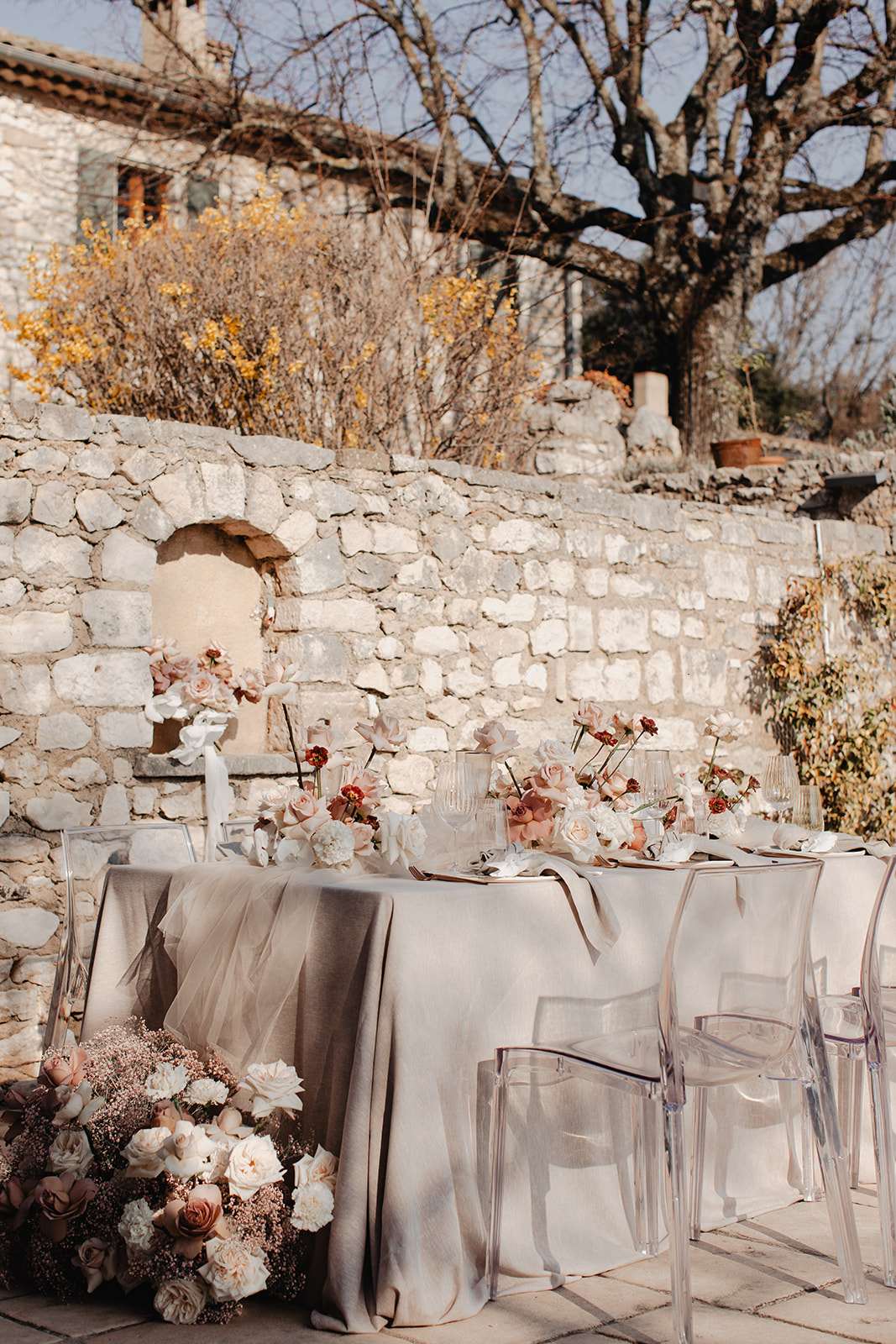 An outdoor reception table setup photographed in a courtyard against a rustic stone wall, with no people present. The long rectangular table is dressed in a greige linen tablecloth with a sheer tulle runner draping over the edge, and is surrounded by clear acrylic ghost chairs. The floral design features a cascading arrangement at the table's end and several centerpieces composed of ivory garden roses, blush and caramel-toned roses, deep burgundy ranunculus, blush gypsophila, and white magnolia-style blooms on dark stems. Place settings include ribbed glassware, gold flatware, and neutral-toned plates. The overall color palette is warm greige, blush, ivory, and terracotta, with a romantic yet modern aesthetic. This is a medium-wide portrait-orientation shot capturing the full length of the table.