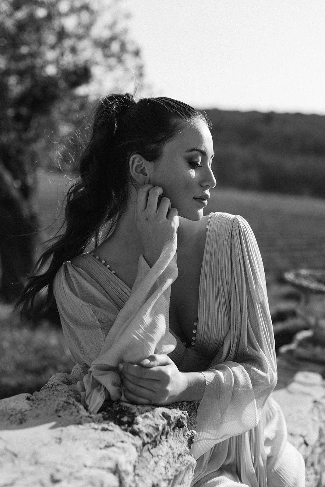 Bride in pleated-sleeve gown seated on rocky surface with pearl necklace in dramatic B&W sunlight