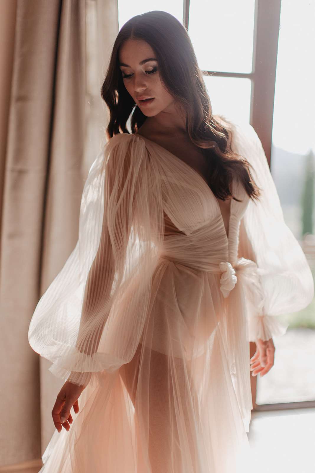 A close-up portrait of a bride standing near a large window during a getting-ready moment, shot indoors with natural backlight streaming through sheer curtains. She wears a blush-ivory tulle wedding gown with dramatically voluminous bishop sleeves featuring fine pleated detailing and a deep V-neckline with a wrapped bodice. The skirt is soft tulle in a pale blush tone, and a small floral wrist detail is visible at the cuff. Her dark brown hair falls in loose waves over one shoulder, and her gaze is directed downward. The composition is a medium-close portrait with a shallow depth of field, emphasizing the texture of the dress fabric against the soft, warm light of the window.