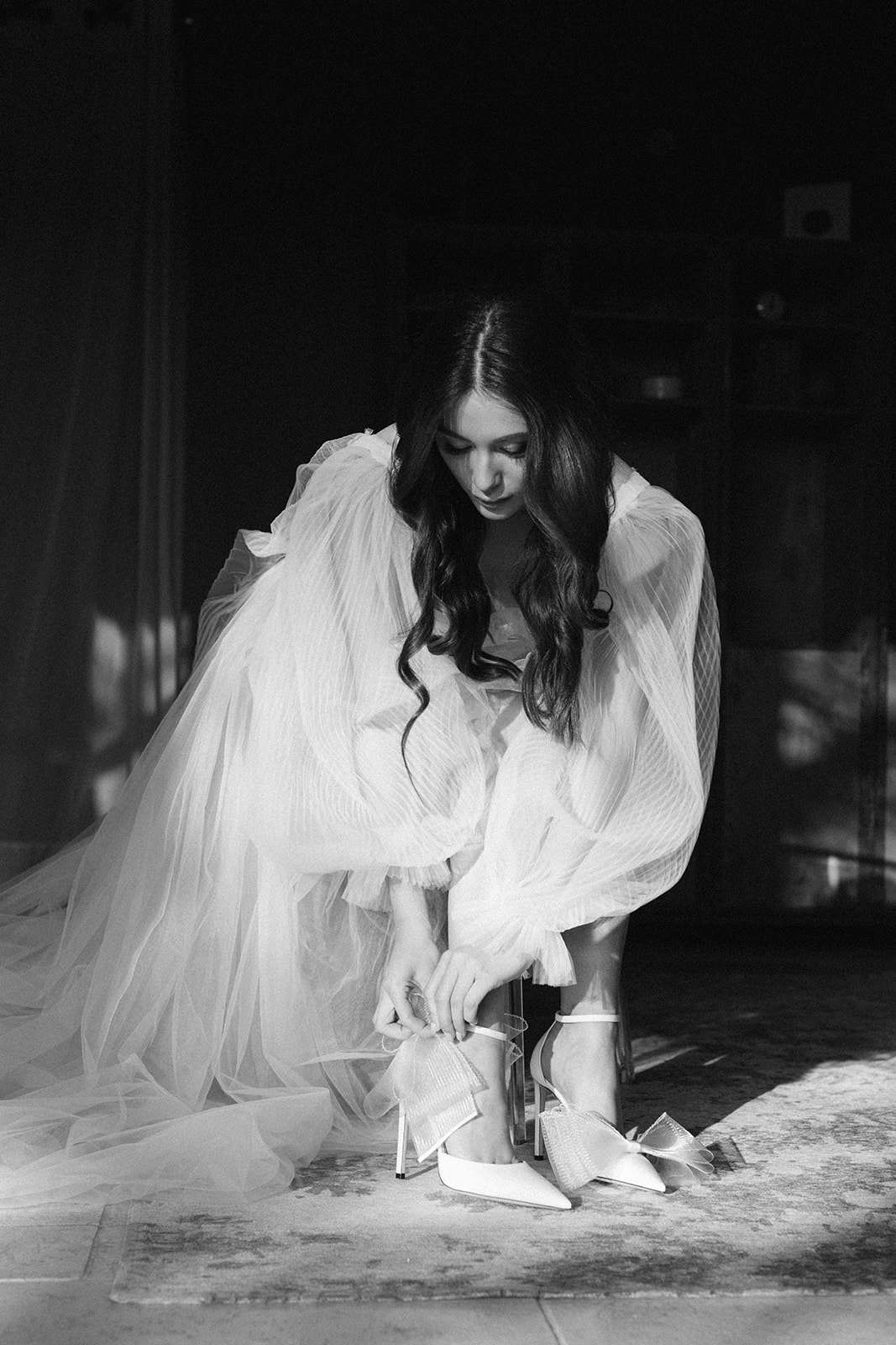 Black and white portrait of bride in sheer pleated robe fastening strappy heeled shoes in dramatic window light