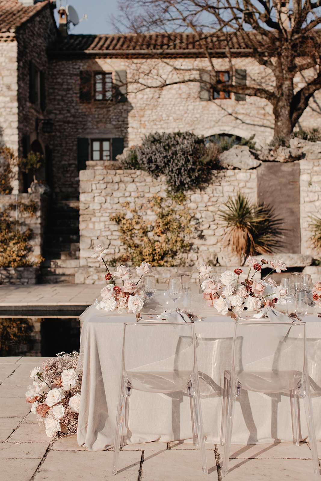 An outdoor reception table setup photographed in a medium-wide shot against the backdrop of a rustic stone mas or bastide with terracotta roof tiles. The rectangular dining table is dressed with a pale greige linen tablecloth and set with clear glassware, white folded napkins, and gold cutlery. Transparent acrylic ghost chairs line the visible side of the table. The floral styling runs as a low garland along the tabletop and spills onto the floor at one end, featuring blush roses, ivory peonies or ranunculus, dusty pink cosmos-style blooms, burgundy accent flowers, and dried gypsophila in soft neutral and muted rose tones. The table appears positioned beside a dark reflecting pool or water feature on a stone-paved terrace. The overall decor palette is neutral and muted — greige, blush, ivory, and warm terracotta — with a modern-classic styling approach mixing natural textures with contemporary acrylic furniture. Potential venue feature image.