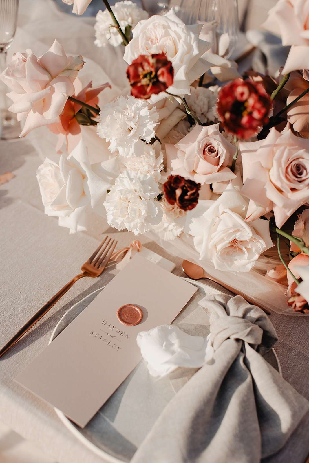 A close-up detail shot of a wedding reception place setting for a couple named Hayden and Stanley. The table is dressed with a light grey linen tablecloth and a loosely knotted grey linen napkin, with rose gold flatware including a fork and knife visible. A beige card printed with the couple's names features a rose gold wax seal as a menu or place card. The centerpiece arrangement spills across the table and includes blush roses, white carnations, ivory garden roses, and deep burgundy ranunculus or poppies, creating a soft romantic palette with rich contrast accents. The overall styling theme is modern classic with a warm rose gold, blush, and grey color palette.