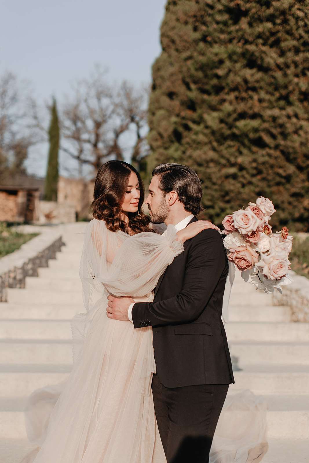 A couple portrait taken outdoors on wide stone steps, likely at a French château or domaine. The bride wears a blush-toned tulle gown with sheer, flowing long sleeves and a deep V-neckline, her dark wavy hair styled down with a small dark hair accessory; she holds a rounded bouquet of blush and dusty rose garden roses with ivory blooms. The groom wears a black suit with a white shirt and no tie, his dark hair slicked back. The two face each other closely in an embrace, with the bride's arm around the groom's shoulder. The styling is modern and romantic with a soft, neutral color palette. The shot is a mid-length portrait with warm natural light and a shallow depth of field.