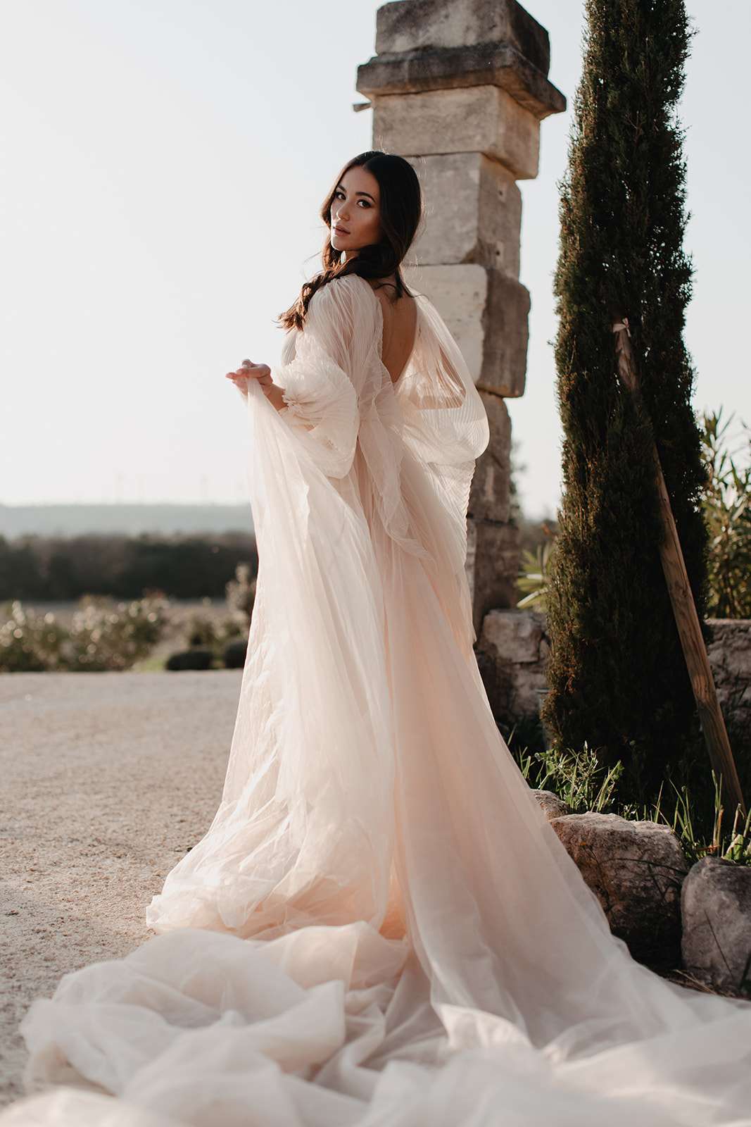 Bride in blush pink gown with bishop sleeves glancing over her shoulder on a gravel terrace beside a stone column