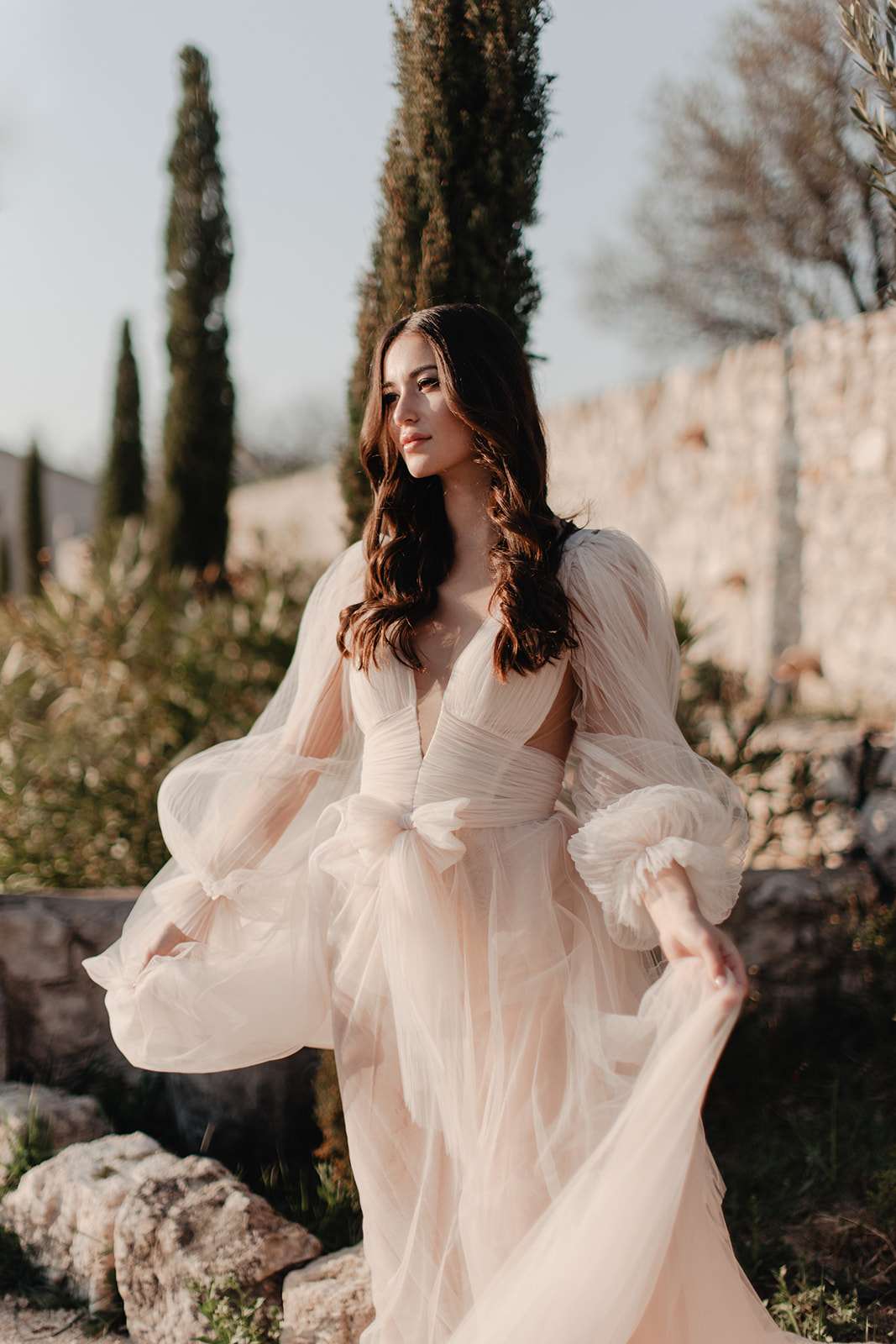 Bride in blush tulle gown with bishop sleeves holding her skirt on the grounds of a stone property with cypress trees
