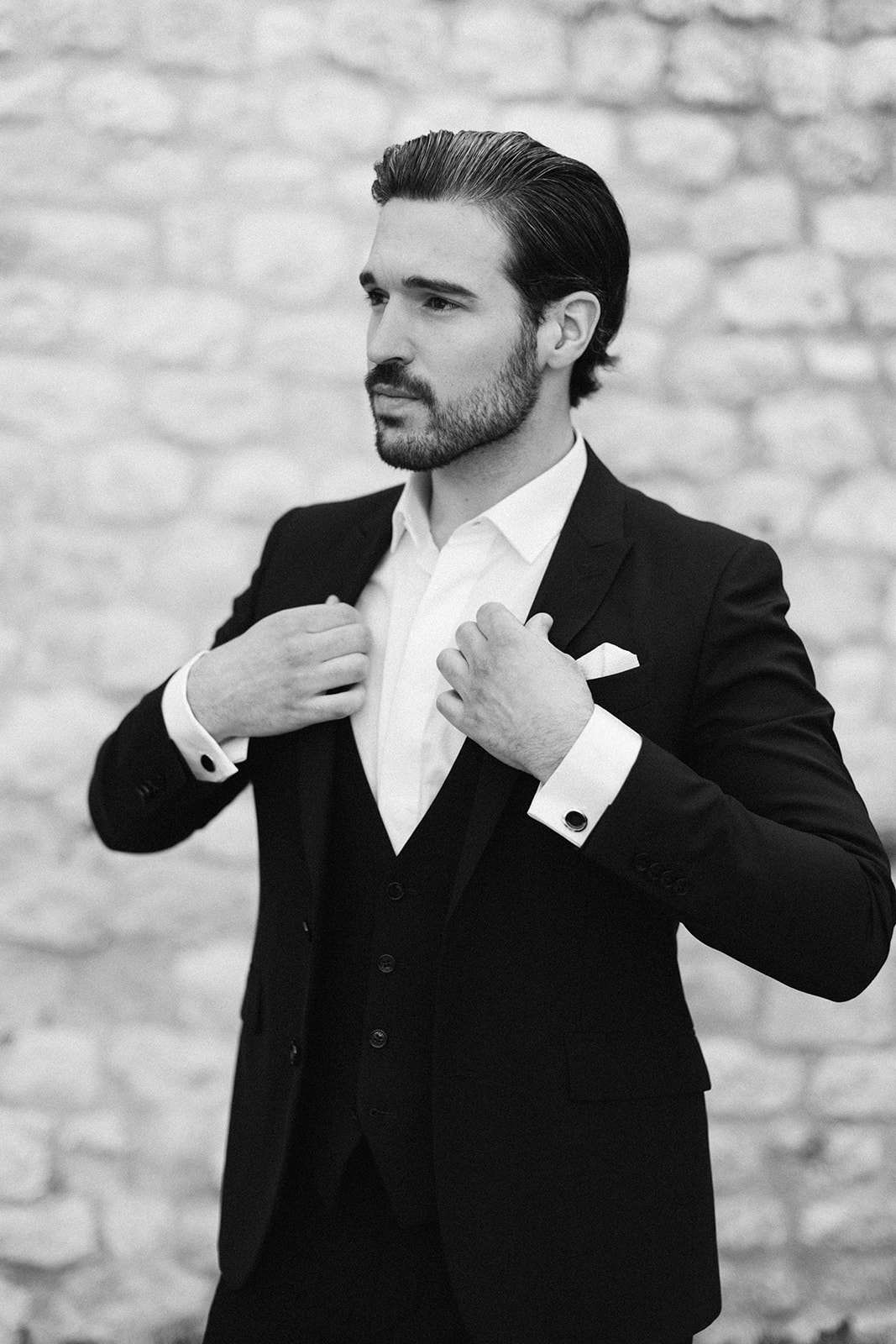 Black and white portrait of groom adjusting collar wearing three-piece suit with pocket square