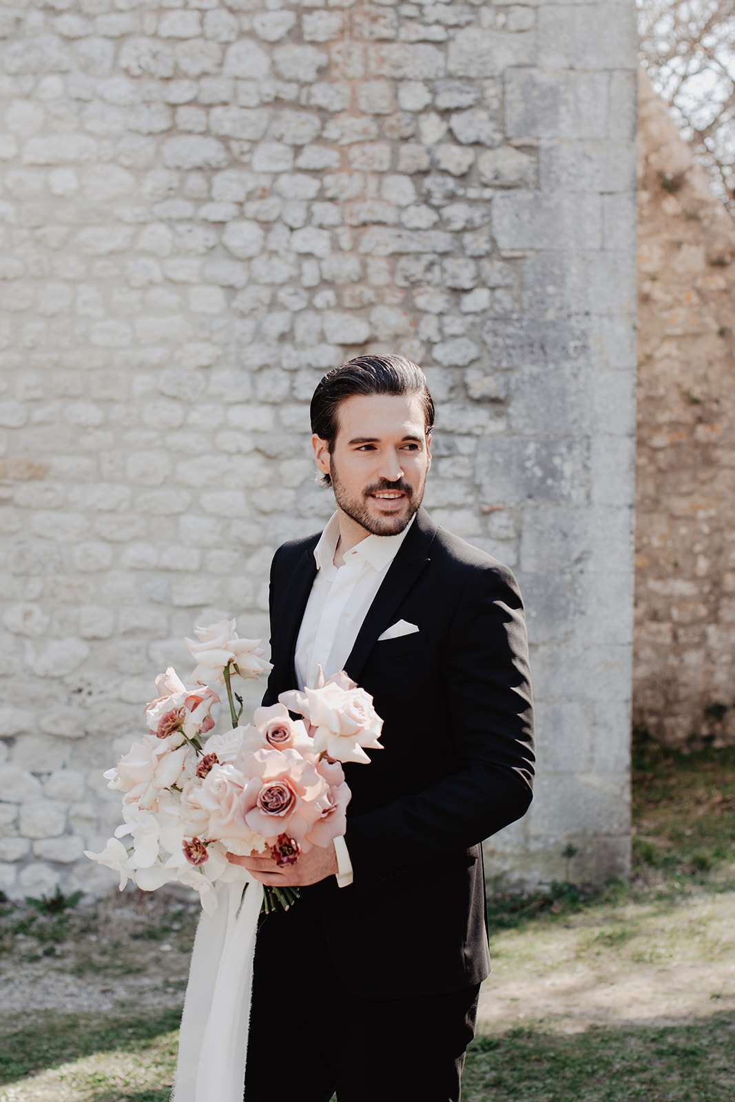 Groom in black suit holding bouquet of blush roses, mauve garden roses, and white orchids