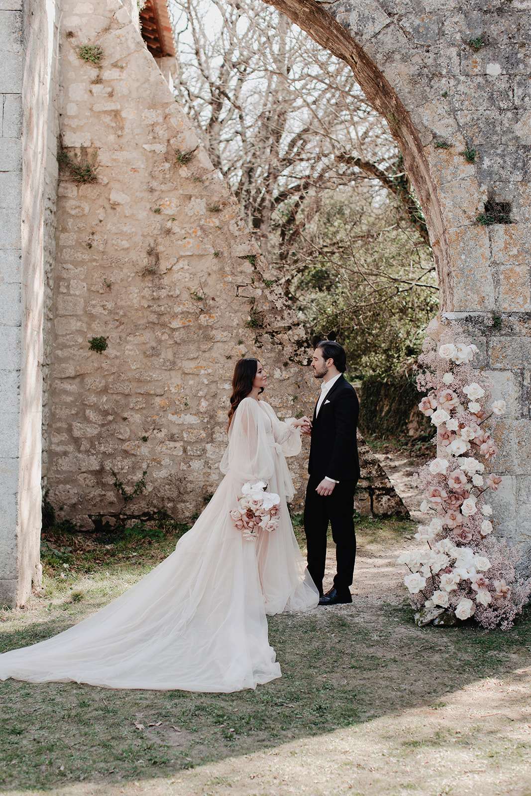 Bride and groom holding hands under stone ruin arch beside tall blush and white rose floral column
