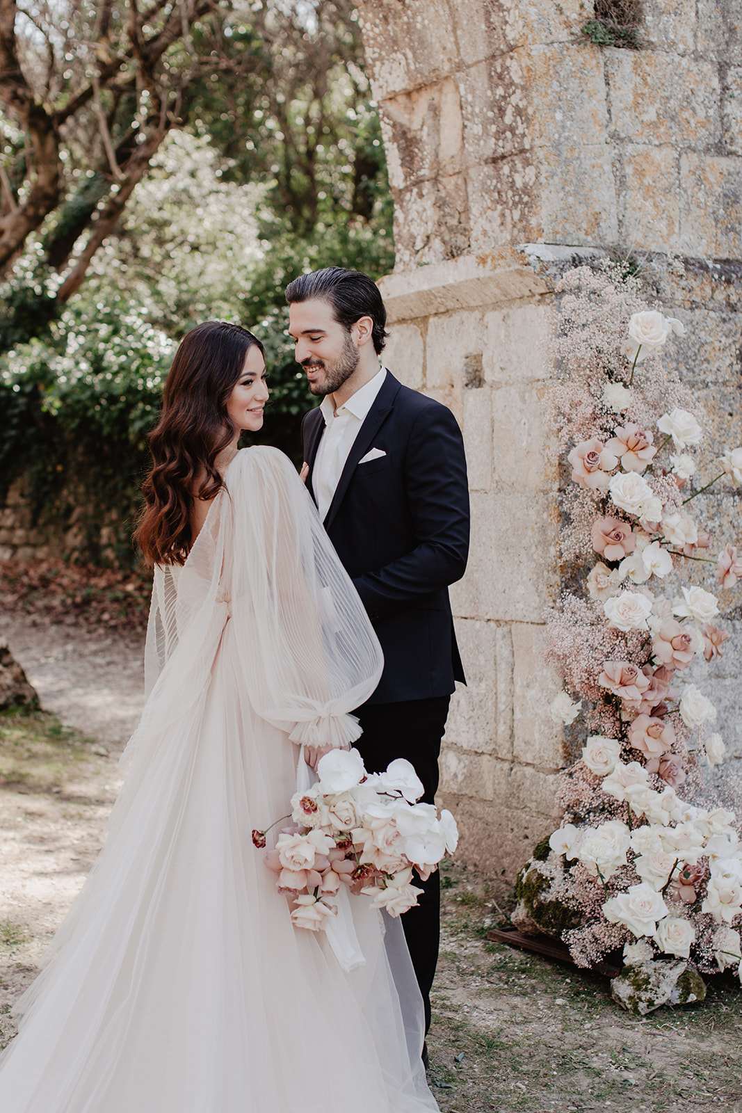Couple before stone wall with vertical blush rose column and bride holding blush and orchid bouquet