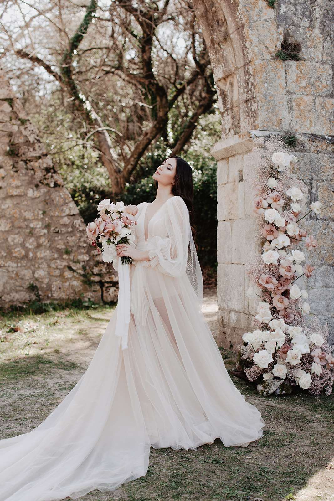 A bridal portrait shot outdoors at a historic stone ruin or chapel, with the bride posed in a three-quarter-length stance against aged limestone masonry. She wears a semi-sheer ivory tulle gown with billowing long sleeves, a deep V-neckline, and a sweeping train, styled in a romantic boho aesthetic. She holds a loose bouquet of blush and dusty rose garden roses, pale pink magnolias, and cream blooms with long trailing ribbon. To her right, a stone pillar or column is decorated with a cascading floral installation of ivory and blush roses, dusty mauve garden roses, and dried baby's breath arranged from top to bottom.