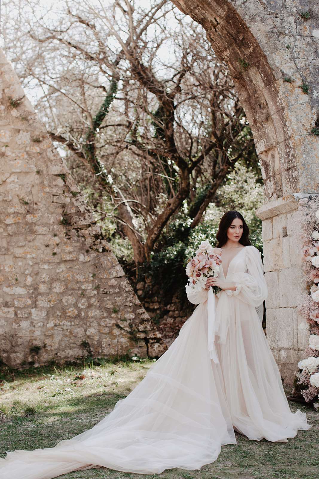 Bride in bishop-sleeve ballgown holding blush rose and orchid bouquet at stone abbey archway