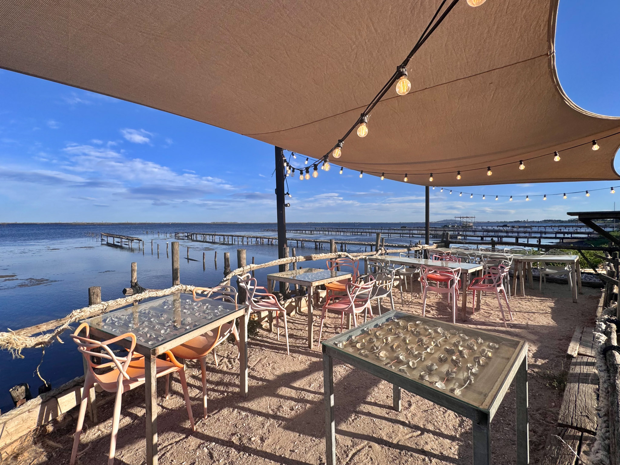 Waterside terrace with coral chairs, glass-topped tables, shade sails, and string lights overlooking oyster lagoon