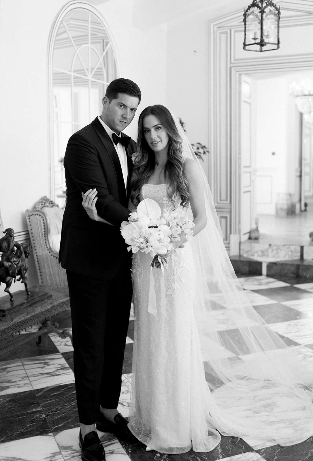 This is a black-and-white couples portrait of a bride and groom posing together indoors in what appears to be a French château or grand manor house. The groom wears a black tuxedo with a bow tie and loafers, while the bride wears a fitted strapless lace gown with floral appliqué detailing and a long cathedral-length veil that trails across the floor. She holds a round bouquet of large blooms that appear to be peonies, orchids, and anthurium-style flowers. The interior setting features a black-and-white checkered marble floor, ornate white molded wall paneling, an arched mirror, a bronze equestrian sculpture on a gilt console table, and a wrought-iron lantern pendant light visible in the background. The composition is a medium full-length portrait with high contrast tones, shot in a classic editorial style.
