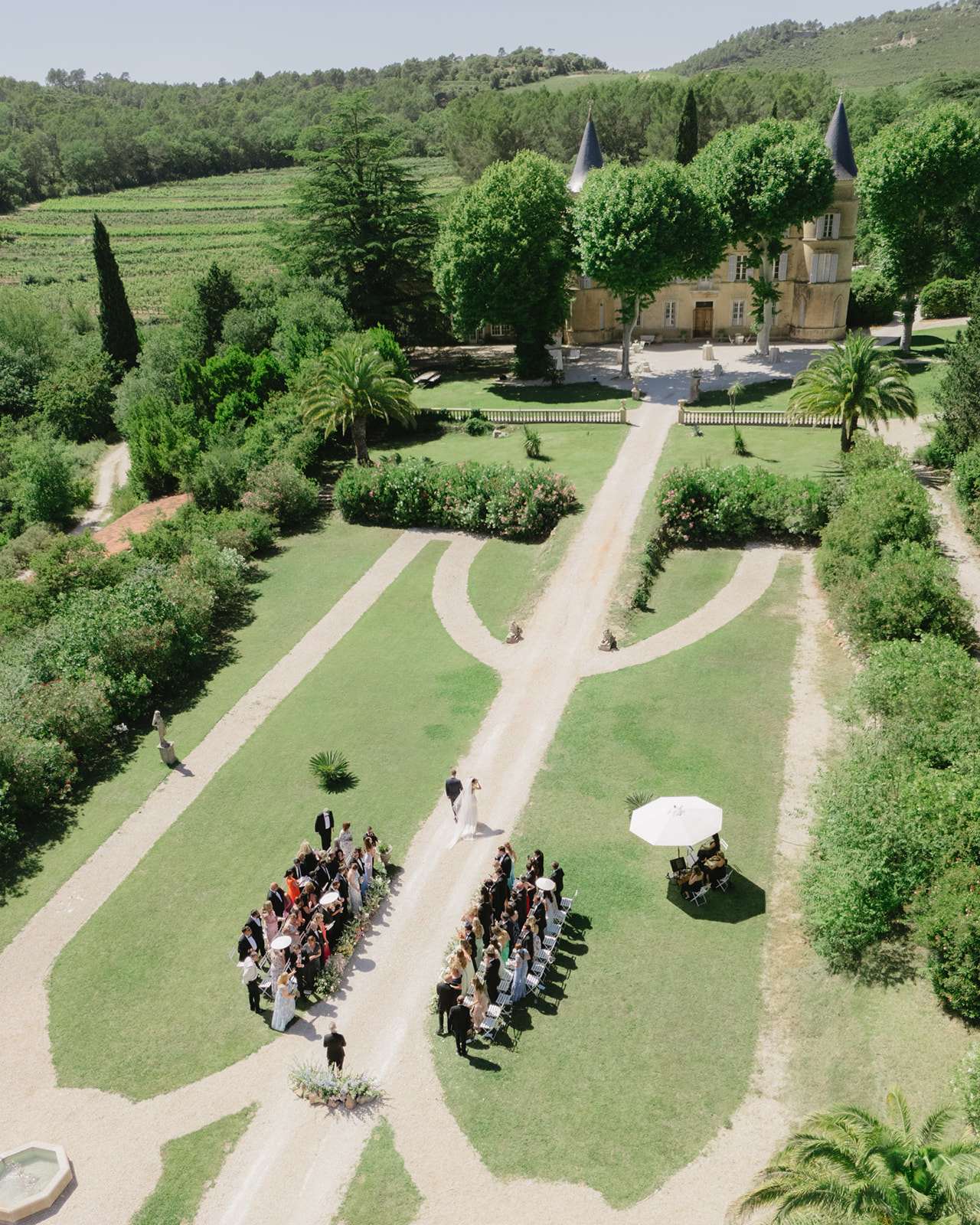 Aerial view of outdoor wedding ceremony on chateau allee with guests seated in white chairs and vineyards beyond