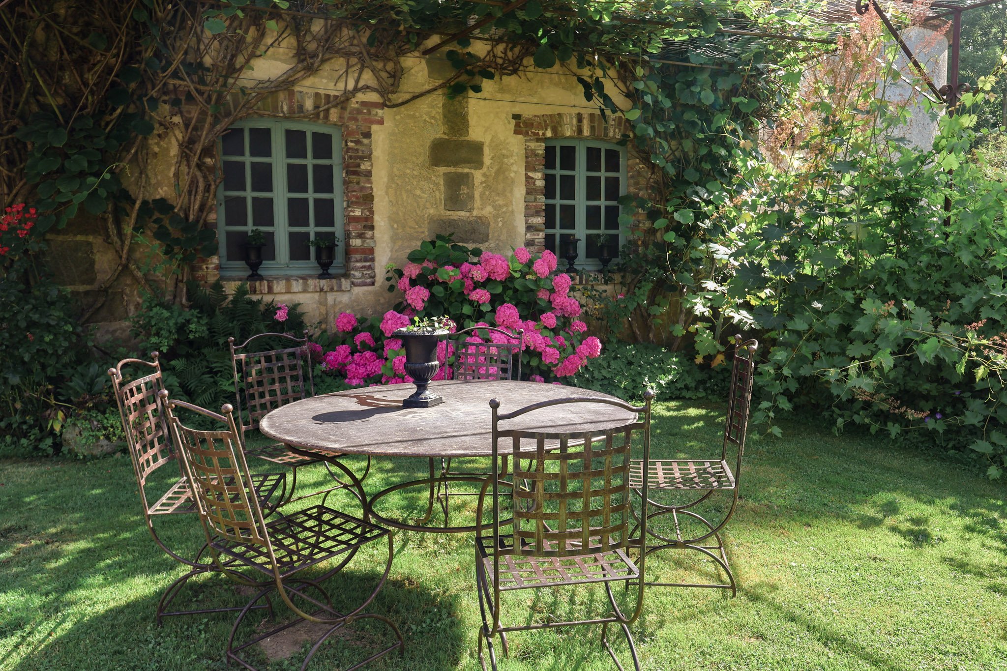 Round stone table with wrought-iron chairs on lawn beside hot pink hydrangeas at a rustic French country property