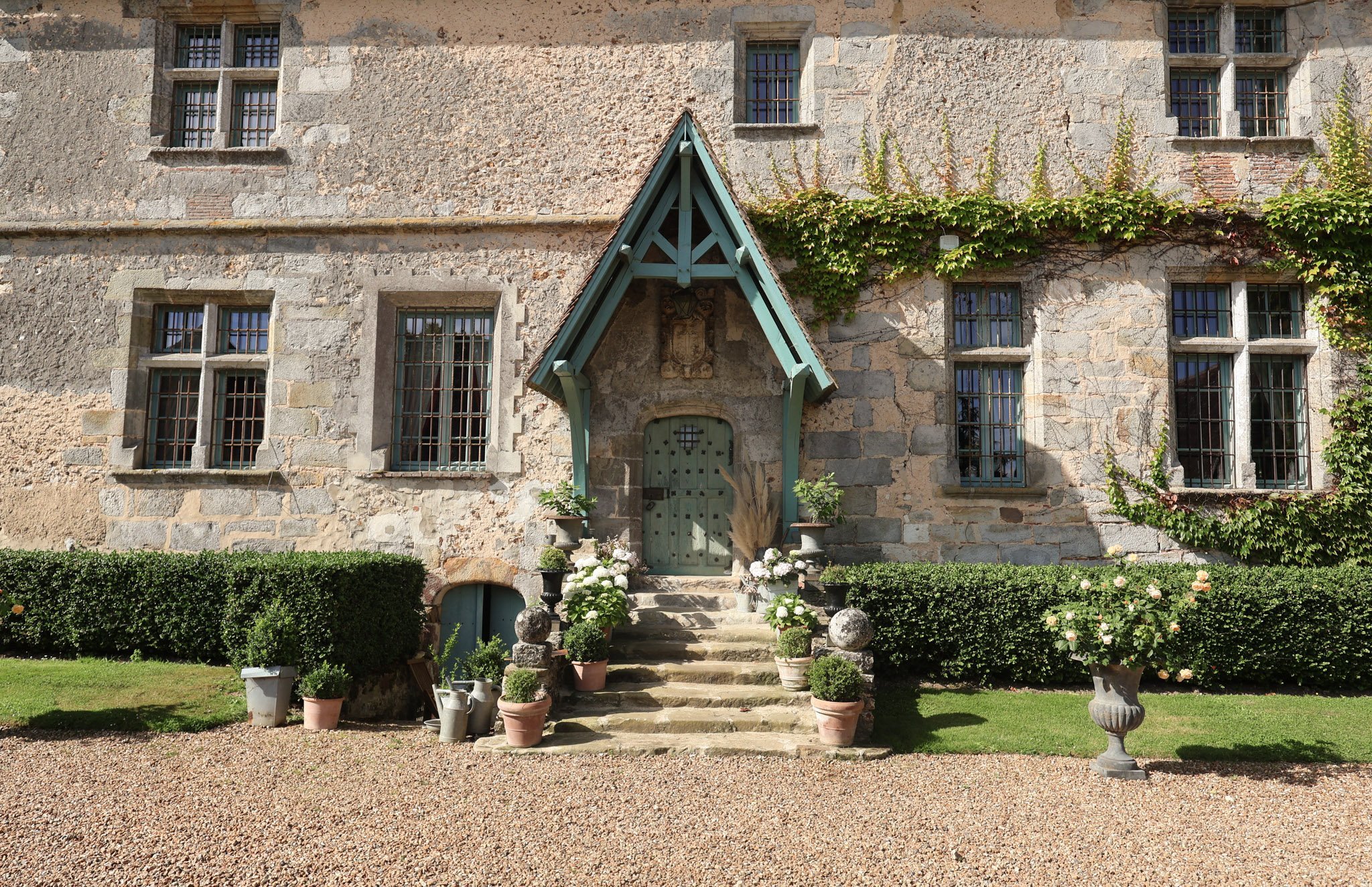 French chateau entrance with teal-painted door and porch canopy, flanked by topiary and hydrangeas on gravel path
