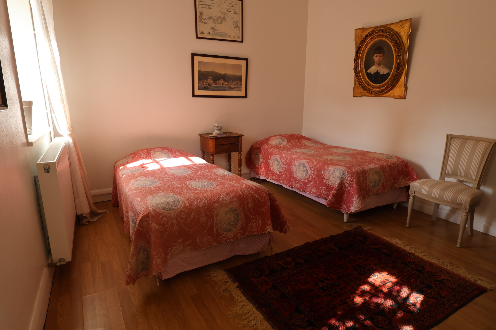 Guest bedroom with two single beds in red and ivory bedspreads, antique furniture, and framed artwork in French manor house