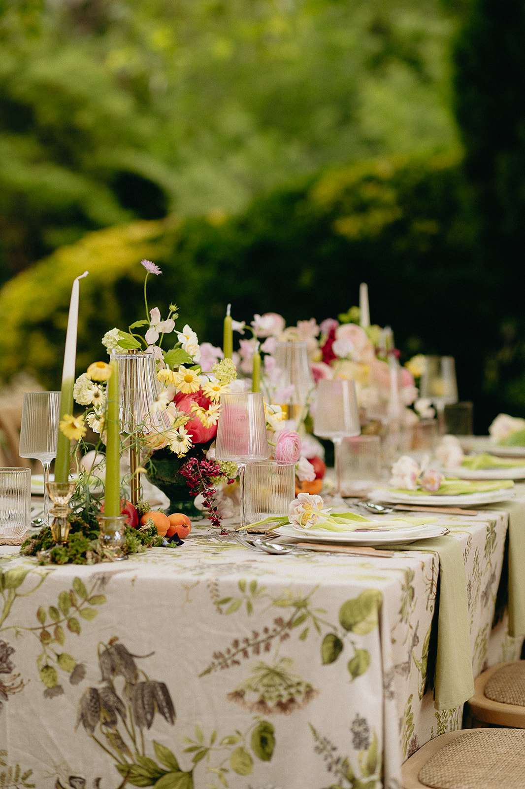 A detailed close-up of an outdoor reception tablescape set in a garden setting with no guests present. The long dining table is covered in a botanical-print linen tablecloth featuring green and brown leaf and fruit motifs on an ivory background. Centerpieces consist of lush floral arrangements including yellow daisies, pink ranunculus, coral peonies, burgundy berries, and small white blooms, accented with moss and scattered apricots at the base. Sage green taper candles are placed in gold candleholders throughout the table runner arrangement. Place settings include white ceramic plates layered with gold chargers, lime green folded napkins, silver cutlery, and individual flower buds as napkin accents. Glassware includes ribbed clear highball glasses, pink-tinted wine glasses, and amber-toned tumblers. The overall styling palette combines botanical greens, warm corals, soft pinks, and gold, with a garden-party aesthetic.