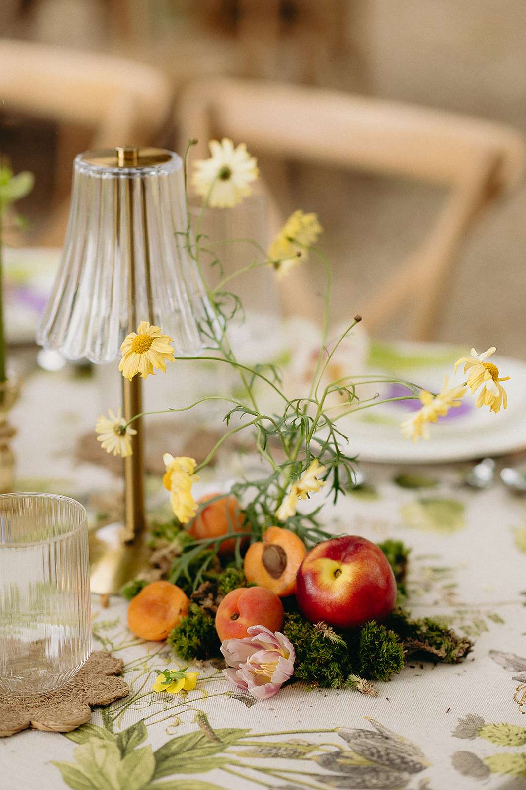 Close-up of reception centerpiece with nectarines, chamomile flowers, pink tulip, and brass lamp on botanical linen