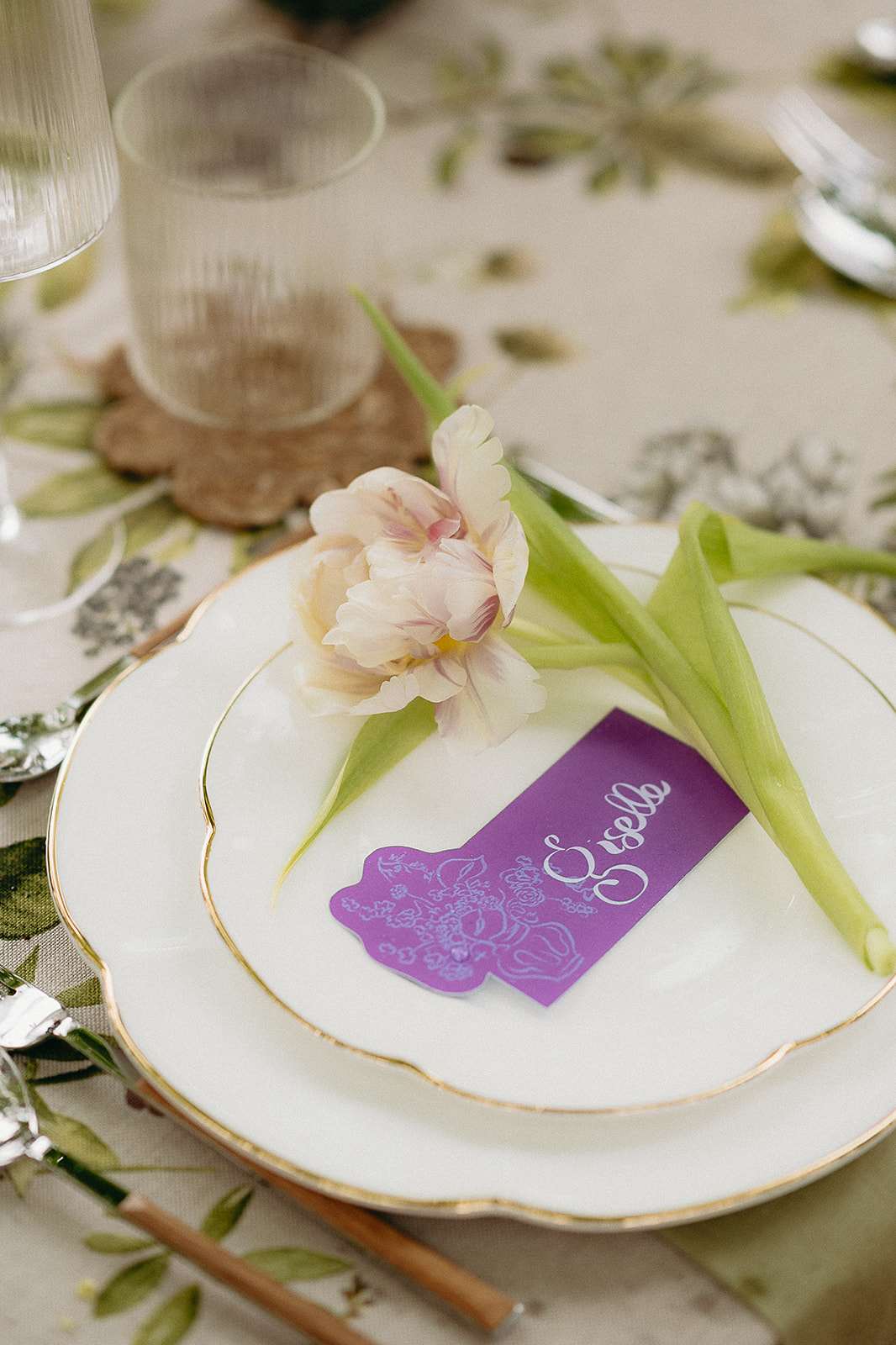 Place setting with gold-rimmed plates parrot tulip and purple calligraphy card on botanical linen