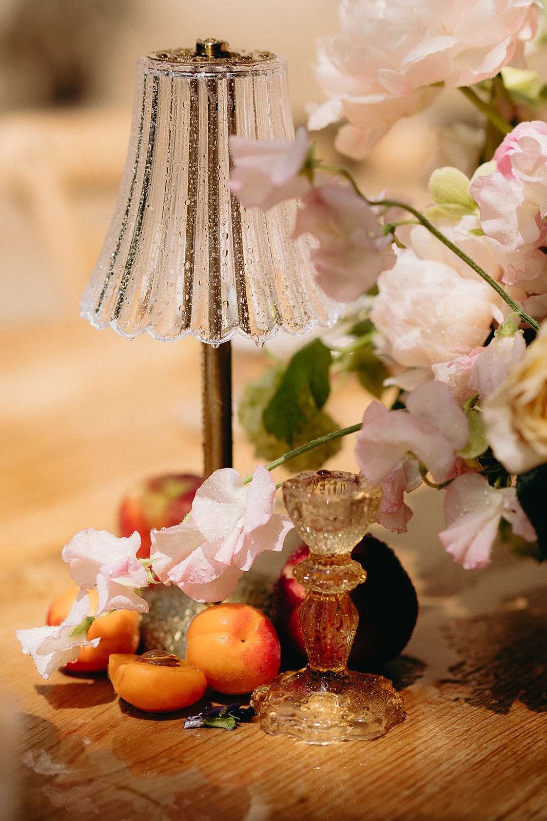 A close-up detail shot of a wedding reception tablescape styled on a raw oak wood surface. The arrangement features a small glass table lamp with a ribbed, scallop-edged shade on a brass stem, accompanied by an amber pressed-glass candleholder. Fresh apricots and peaches — some halved — are scattered across the table alongside loose blush sweet peas and small dark plums, creating an organic, garden-harvest aesthetic. A lush floral arrangement of blush peonies, soft pink sweet peas, and cream garden roses fills the upper right of the frame, and all elements show water droplets suggesting an outdoor or recently misted setting. The overall decor palette is warm peach, blush pink, and amber, reflecting a romantic French countryside or organic maximalist styling approach.