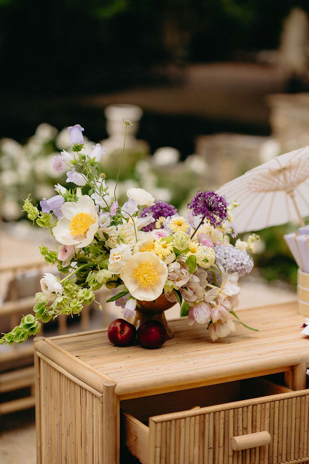 A close-up detail shot of a floral arrangement displayed on a rattan side table with an open drawer, set outdoors at what appears to be a cocktail hour or reception. The arrangement is placed in a small bronze-toned pedestal vase and features cream peonies with yellow centers, lavender campanula bell flowers, purple allium globes, pale yellow daisies, soft pink ranunculus, blue-tinted hydrangea, green hops or bells-of-Ireland foliage, and blush alstroemeria. Three deep red plums or nectarines are placed at the base of the vase as a decorative accent. The overall palette is garden-fresh and painterly, mixing purples, creams, yellows, and greens in a loose, organic bohemian style. A cream paper parasol is partially visible in the background, along with additional white floral arrangements and rattan furniture suggesting a cohesive natural, boho-garden styling theme.
