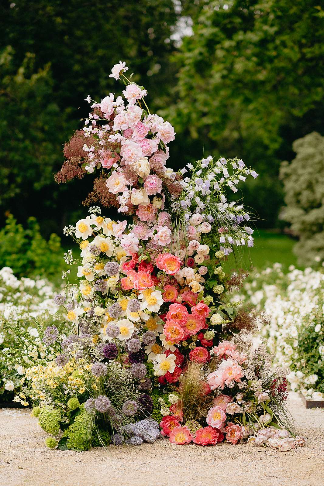 A large-scale freestanding floral installation photographed outdoors on a gravel path, likely used as a ceremony backdrop or decorative focal point. The arrangement rises vertically in an asymmetric, organic shape and incorporates a wide variety of flower types including peonies in coral, pink, and blush tones, cream and yellow garden roses, ranunculus, alliums in purple and mauve, white cosmos-style blooms, lavender bellflowers, viburnum snowball clusters in chartreuse, and dried pampas-style grasses. The color palette transitions from warm coral and yellow tones at the base through mid-range pinks to soft blush and pale pink at the top, with purple and lilac accents extending laterally. Loose wildflower-style stems and textural foliage give the installation a garden-gathered, maximalist aesthetic. The shot is a medium-distance portrait-orientation detail image with the installation centered against a softly blurred garden background.
