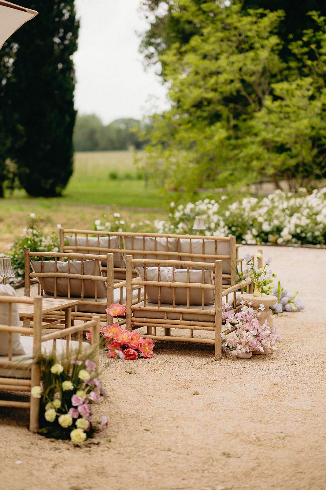 An outdoor cocktail hour or lounge area setup on a gravel surface, with no people present. The seating arrangement features natural bamboo/rattan sofas and chairs with light greige linen cushions, grouped together to create an intimate lounge area. Floral clusters are placed directly on the gravel at the base of the furniture, including coral and pink peonies, soft pink stock flowers, pale yellow flowers, and small arrangements of lavender-blue blooms in low vessels. The styling palette is soft and garden-inspired, combining natural wood tones with pastel florals in a relaxed, organic manner. A wide shot taken from a slightly elevated angle shows the full lounge arrangement with a lush garden border of white flowering shrubs visible in the background.