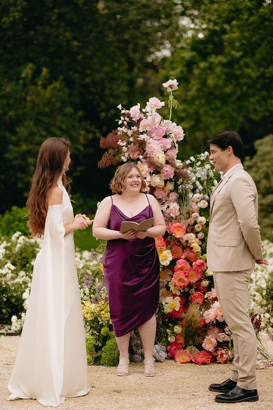 Couple exchanging vows before towering coral, pink, and ivory floral installation in garden ceremony
