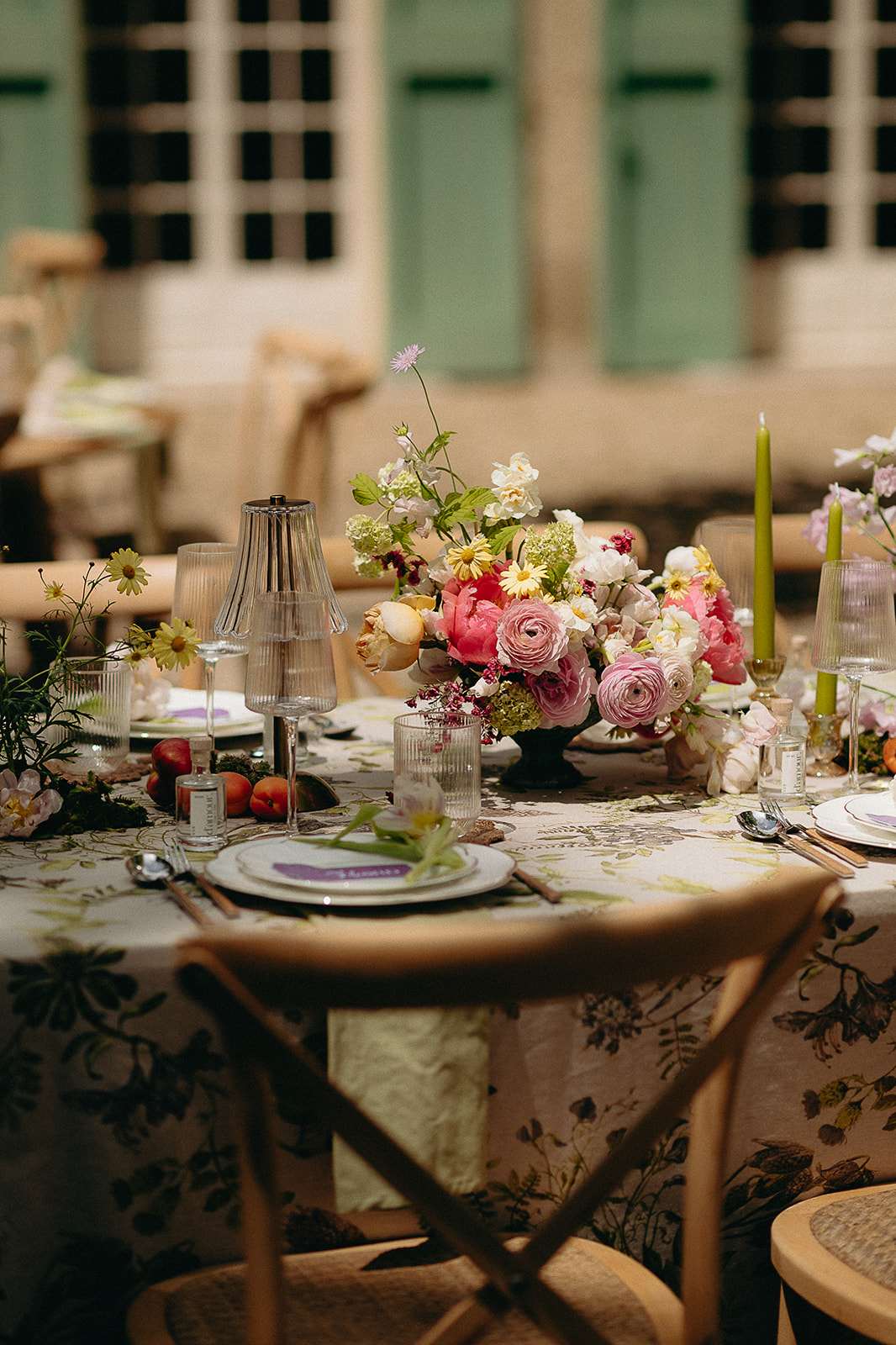 A close-up detail shot of a wedding reception tablescape set outdoors or in a covered courtyard, with sage green shuttered windows visible in the background. The round table is dressed in a botanical-print linen tablecloth featuring dark floral and foliage illustrations on an ivory ground, paired with light wood cross-back chairs. The centerpiece is a low, lush arrangement of coral and hot pink peonies, blush and pink ranunculus, white garden roses, cream hydrangeas, yellow daisy-like blooms, and small wildflowers with loose trailing stems. Olive green taper candles in glass candleholders accent the table alongside ribbed glass tumblers and a small decorative table lamp. Place settings include white porcelain plates stacked on dark charger plates with purple place cards, silver cutlery, and scattered fresh peaches and moss as organic table decorations, giving the overall styling a garden-romantic, maximalist aesthetic.