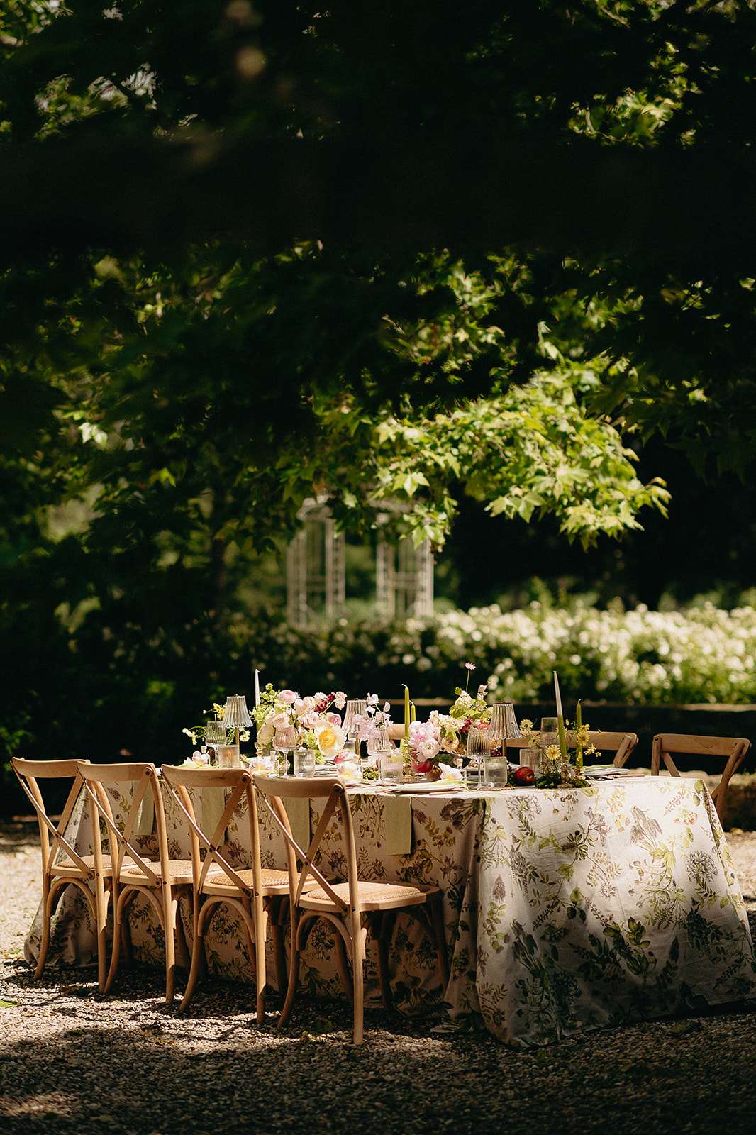 An outdoor reception tablescape set on a gravel surface within a garden setting, with no guests present. The rectangular dining table is covered in a cream botanical-print tablecloth featuring green, yellow, and muted floral and foliage motifs. Natural wood cross-back chairs with rattan seats are arranged along both sides. The centerpiece features low arrangements of blush pink, yellow, and deep pink garden roses and ranunculus, interspersed with small daisy-like flowers and greenery, running the length of the table. Olive green taper candles in ribbed glass candleholders are placed at intervals along the table, alongside clear glassware, plates, and small votive candles. In the background, a white ornamental garden structure or obelisk is partially visible among flowering shrubs and trees. The overall styling is garden-party romantic with a botanical, organic color palette of blush, yellow, green, and ivory. Wide shot with natural dappled light.
