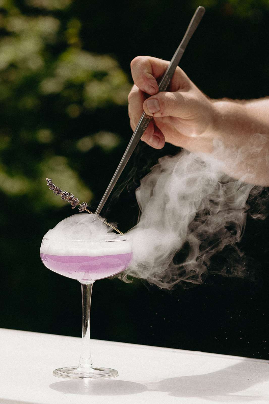 Signature cocktail in coupe glass with lavender garnish and dry ice smoke on white surface
