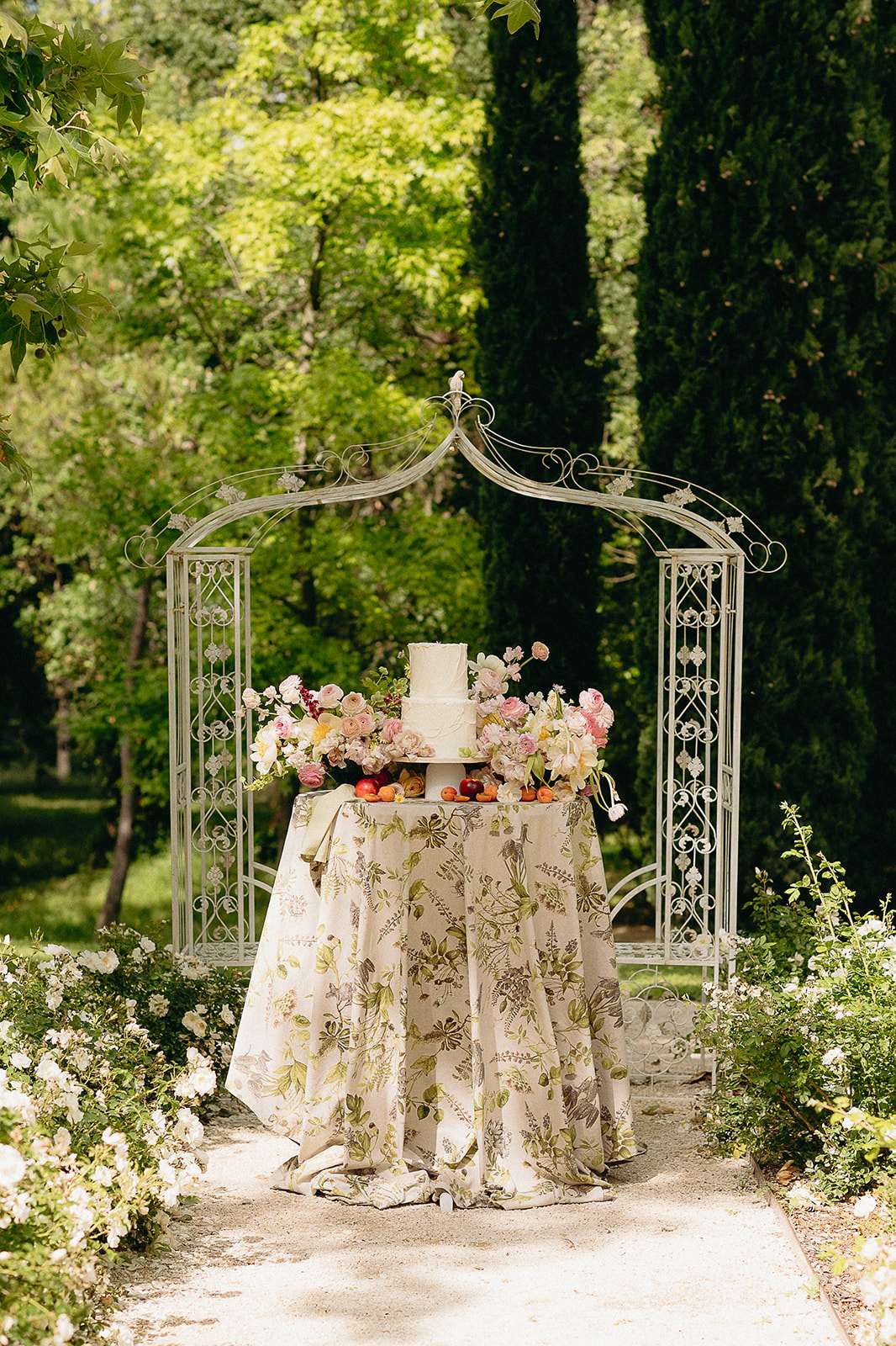 Three-tier white wedding cake on botanical linen under a wrought-iron arch with pink ranunculus and fresh fruit