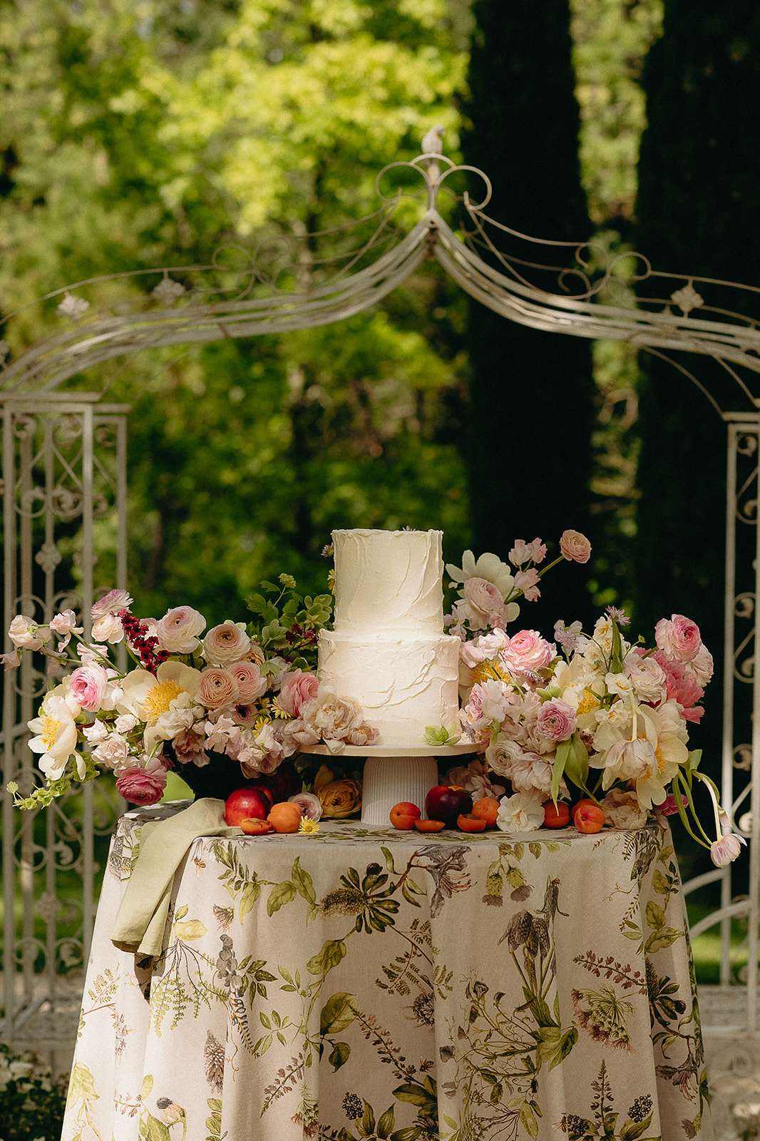 Three-tier ivory buttercream cake on botanical linen with blush ranunculus pink roses scattered fruit and wrought-iron gazebo