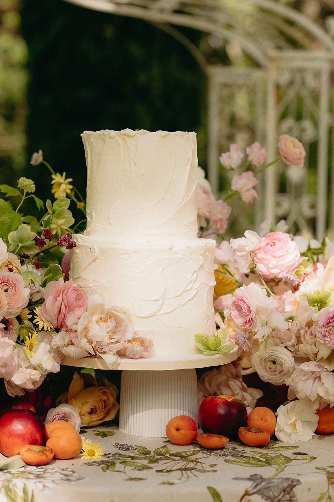 Two-tier ivory buttercream wedding cake on white stand surrounded by blush peonies, ranunculus, and fresh apricots on bota...