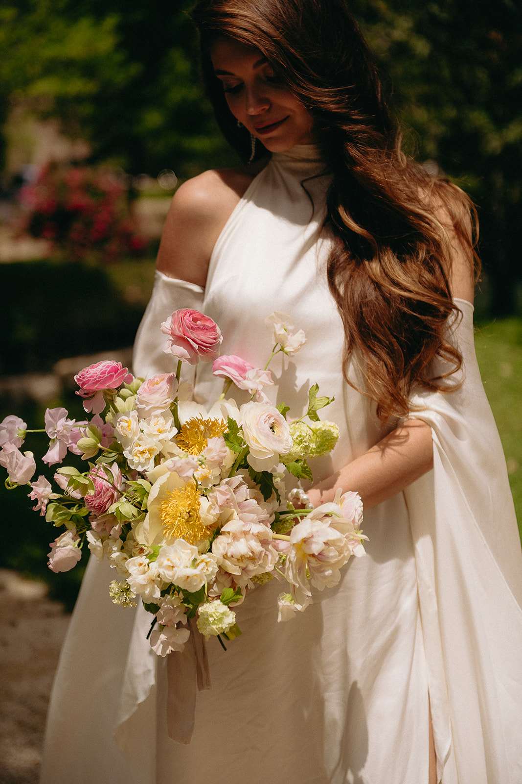 Bride in halter gown with draped cape holding loose bouquet of cream peonies, pink ranunculus, and tulips