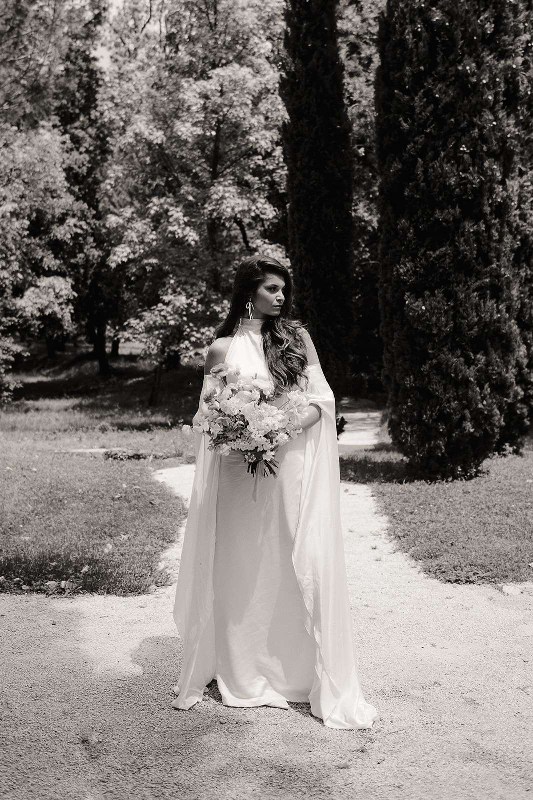 Black and white bride in halter gown with cape sleeves and bouquet on cypress-lined gravel path