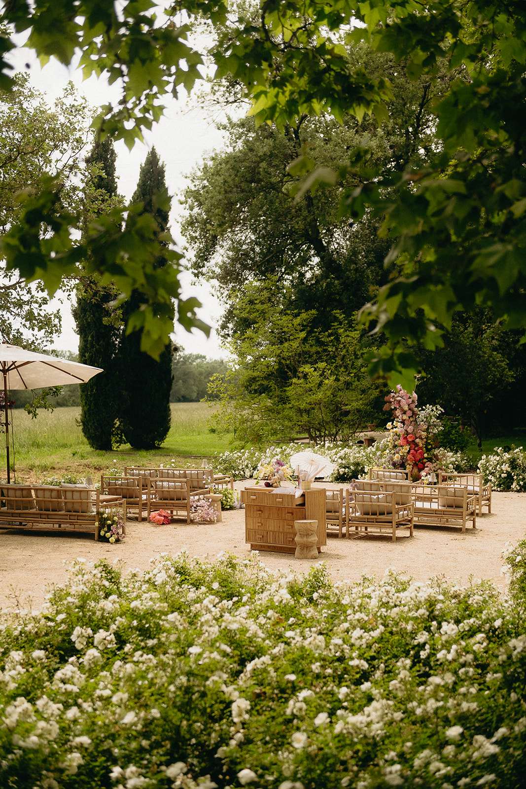 An outdoor wedding ceremony setup photographed from a distance through overhanging tree branches, framing the scene naturally. The seating arrangement features rattan or bamboo-framed sofas and benches arranged in a semicircle around a central wooden altar or officiant's table, styled with a boho-natural aesthetic. A tall floral installation in the right background displays a column of mixed coral, pink, orange, and cream blooms, while loose clusters of colorful flowers are placed on the ground near the seating. A large cream market umbrella is positioned to the left, and the gravel ceremony area is bordered by dense white flowering shrubs in the foreground. Wide shot with no people present, suggesting this is a pre-ceremony setup image.