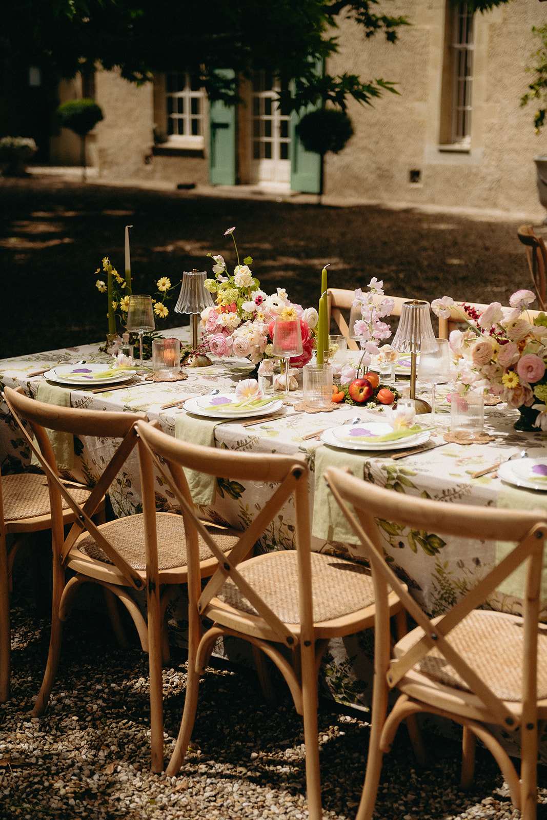 An outdoor reception tablescape shot on a gravel courtyard in front of a French manor house with sage-green painted doors. The long rectangular table is dressed in a botanical-print linen tablecloth featuring illustrated herbs and botanicals, layered with a pale mint green table runner. Place settings include white plates with floral-illustrated chargers, gold flatware, and ribbed glassware in clear and smoky green tones. Centerpieces are lush low arrangements of blush garden roses, hot pink peonies, cream ranunculus, pink sweet peas, and yellow daisy-like blooms, accented with fresh fruit including peaches and tomatoes scattered along the table. Tall olive-green taper candles in brass candlestick holders add vertical interest throughout. Natural wood cross-back chairs with wicker seats surround the table. The overall styling theme is garden-party botanical with a warm, organic color palette of blush, hot pink, cream, and chartreuse green. Wide angled portrait-orientation detail shot with no people present.